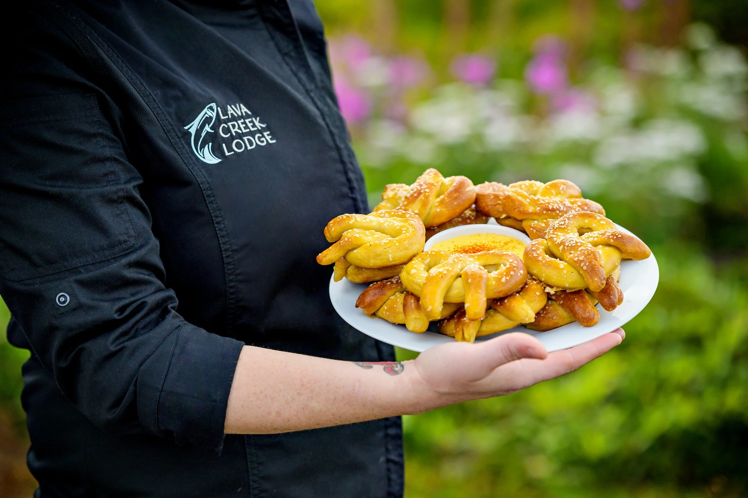 Person wearing a black jacket with 'Lava Creek Lodge' logo holding a plate of pretzels with a bowl of mustard, outdoors with blurred pink and green background.