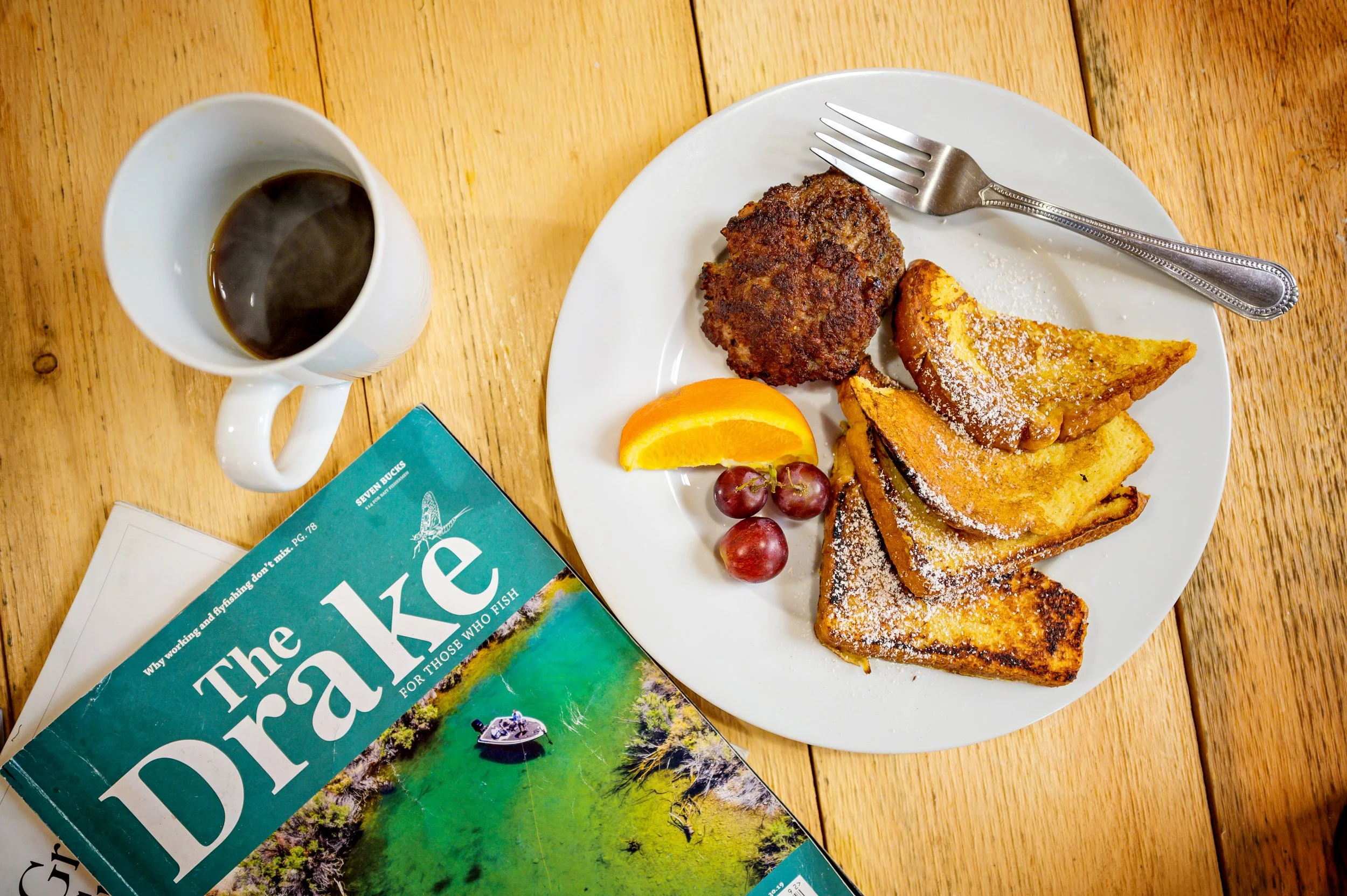 A breakfast plate with French toast dusted with powdered sugar, a sausage patty, orange slices, and grapes. A cup of coffee and a magazine are also on the wooden table.