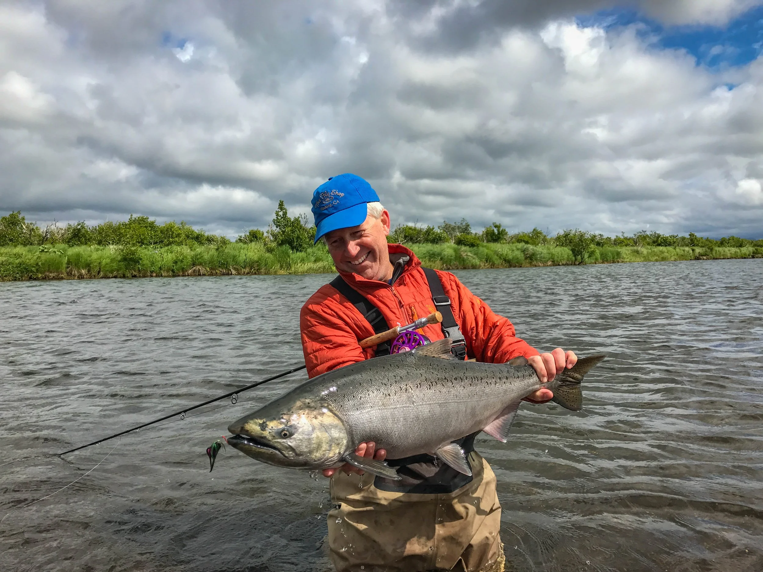 A man smiling while holding a large fish over water, wearing a blue cap, orange jacket, and tan waders, with a green landscape and cloudy sky in the background.