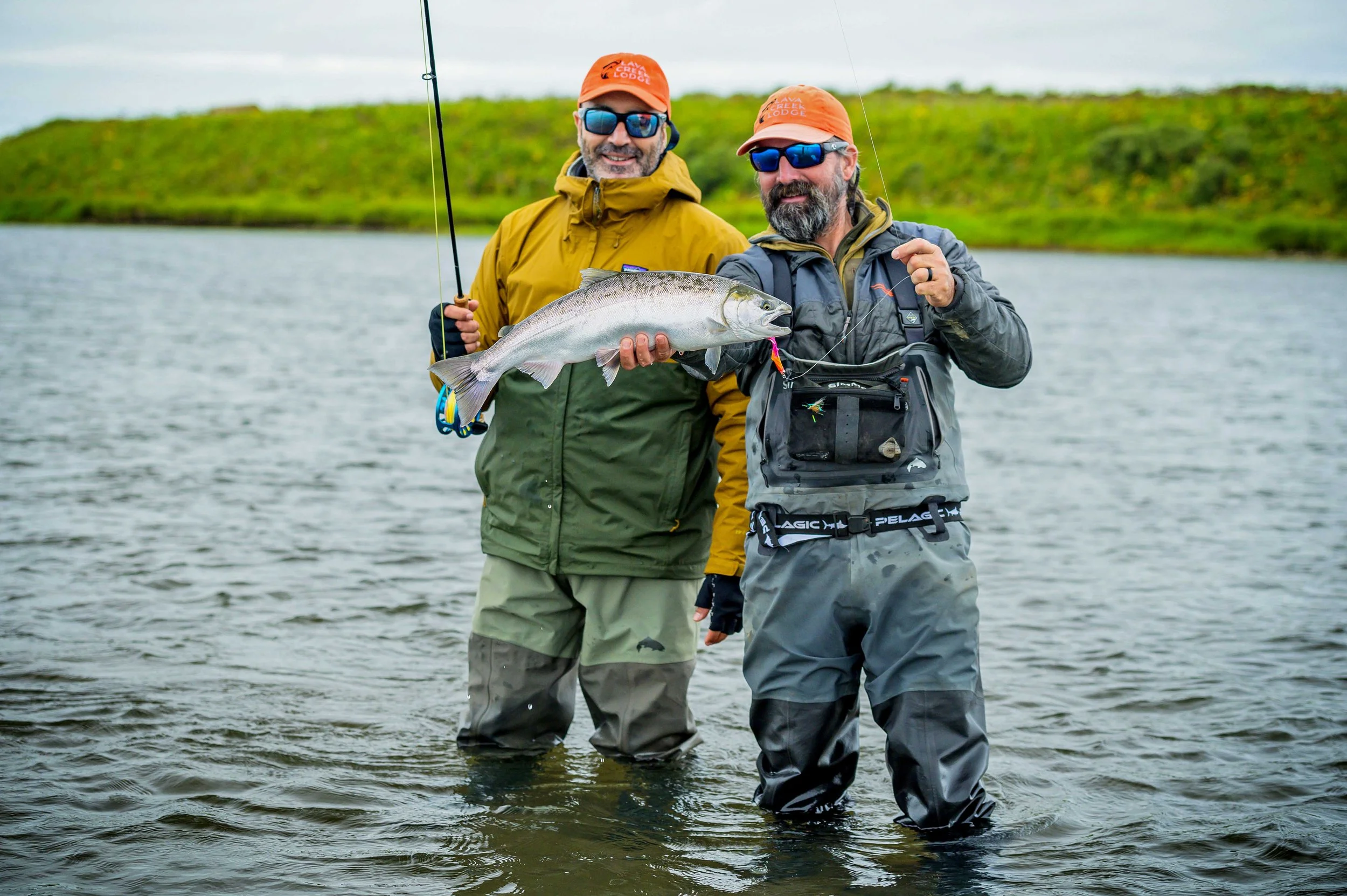 Two men standing in a river, holding a large fish, with a grassy hill in the background. They are dressed in outdoor gear, wearing sunglasses and orange caps, and appear to be fishing.