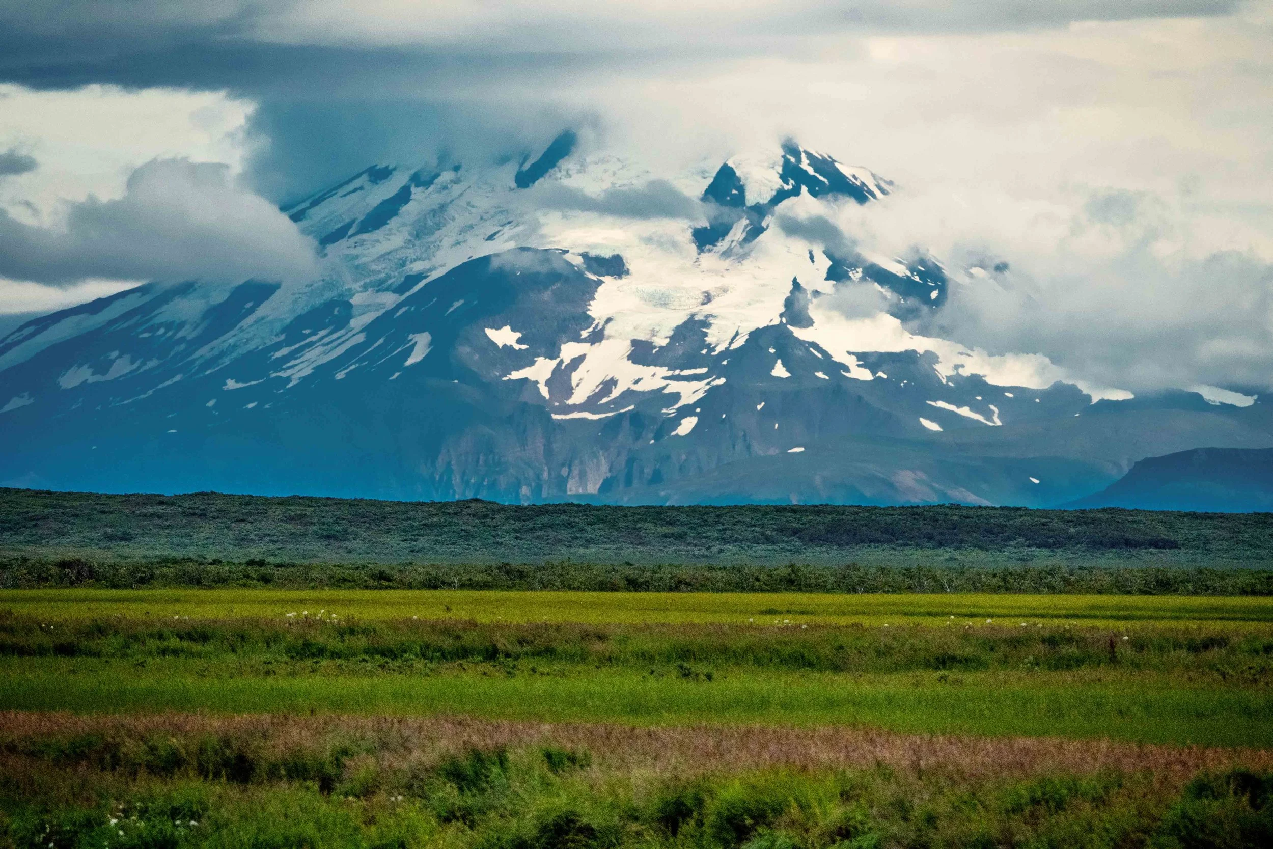 Snow-capped mountain with volcanic features, surrounded by clouds, with green plains in the foreground.