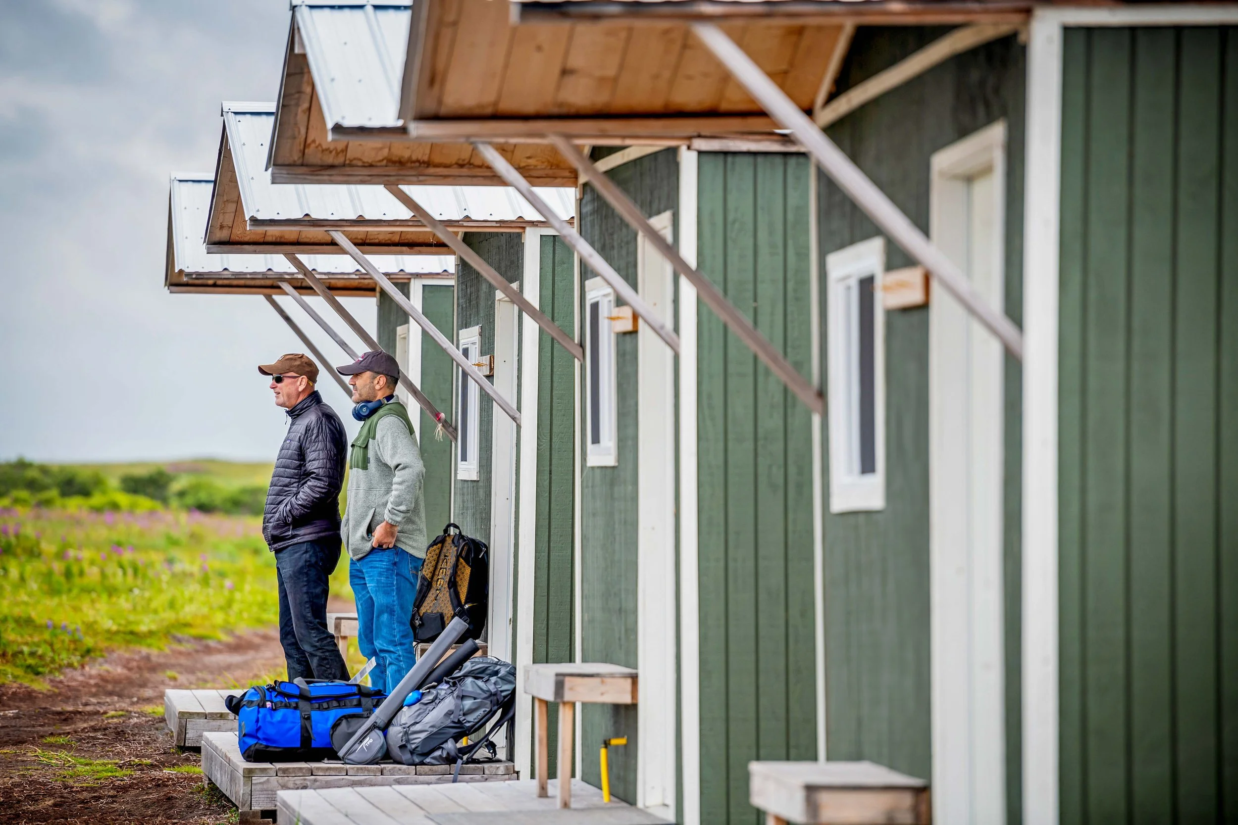 Two men wearing jackets and hats standing outside of green houses on a porch, with backpacks and equipment on the ground, under an overcast sky.