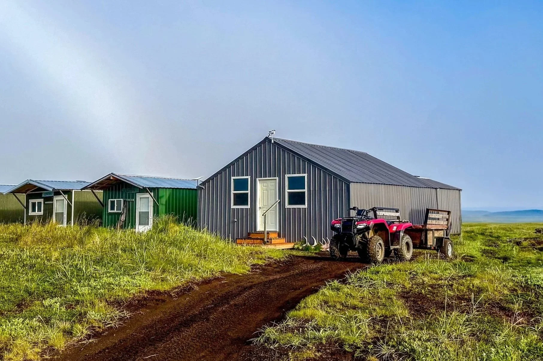 A rural scene featuring a large metal house with a metal roof, two smaller green metal structures to the left, and an ATV with a trailer to the right, all surrounded by green grass under a cloudy sky.