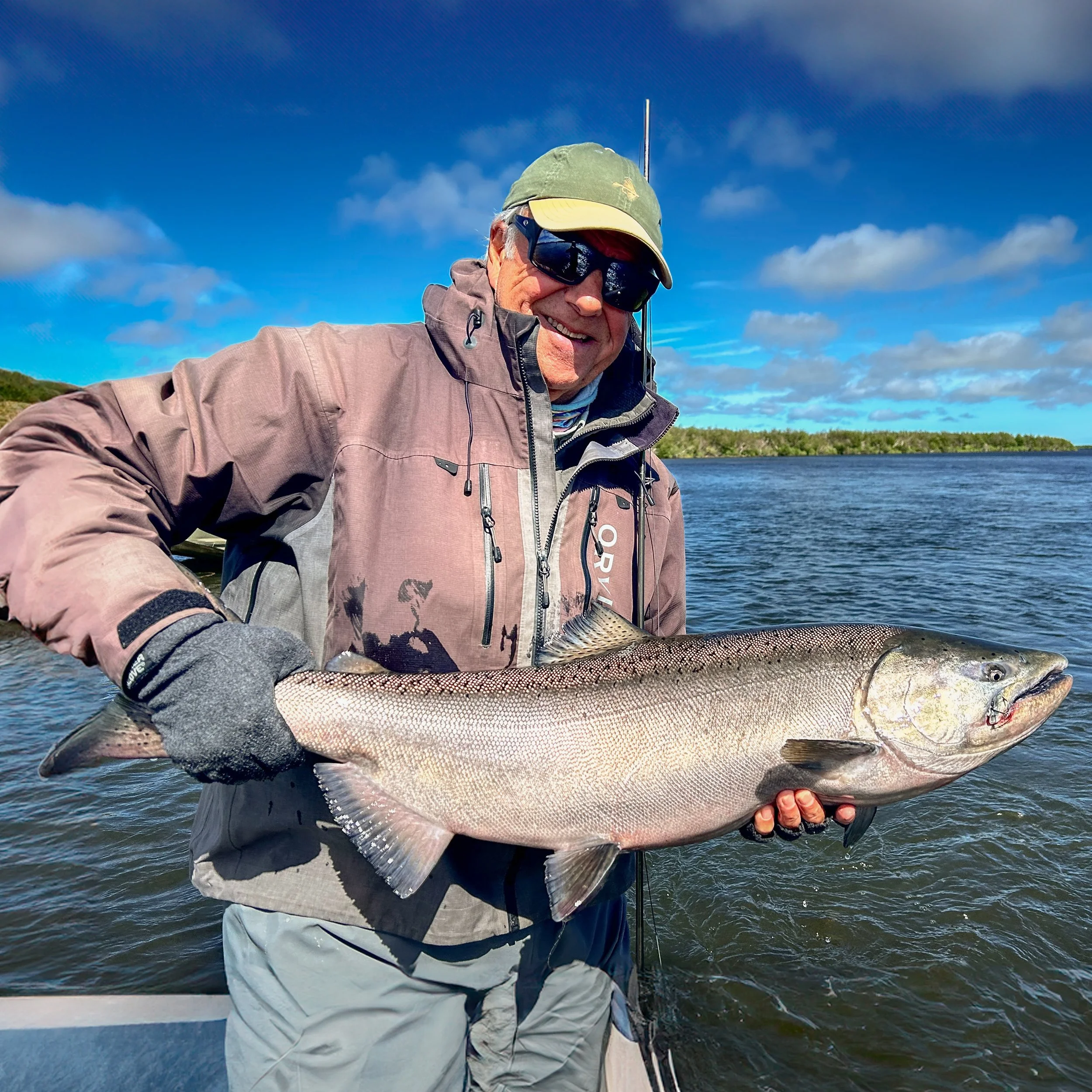 A man wearing a green cap, sunglasses, and a gray jacket holds a large fish he caught in a body of water, with a background of blue sky and clouds.