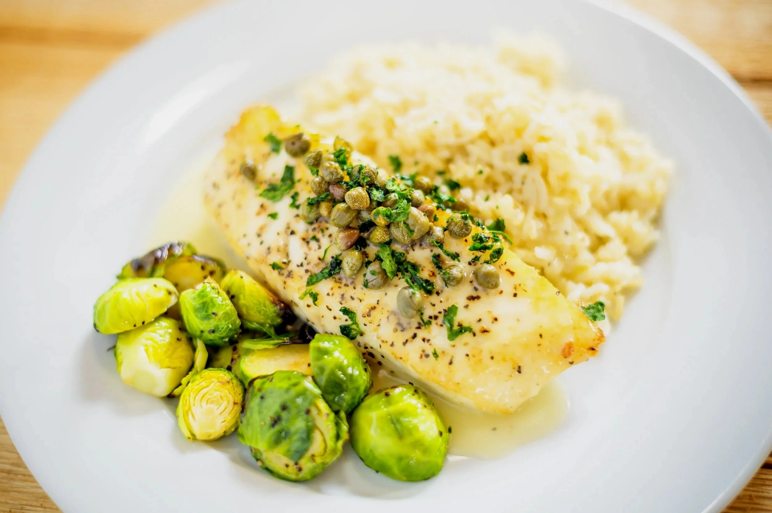 A white plate with a serving of cooked potatoes, garnished with herbs, alongside Brussels sprouts, and a piece of cooked white fish topped with capers and herbs.