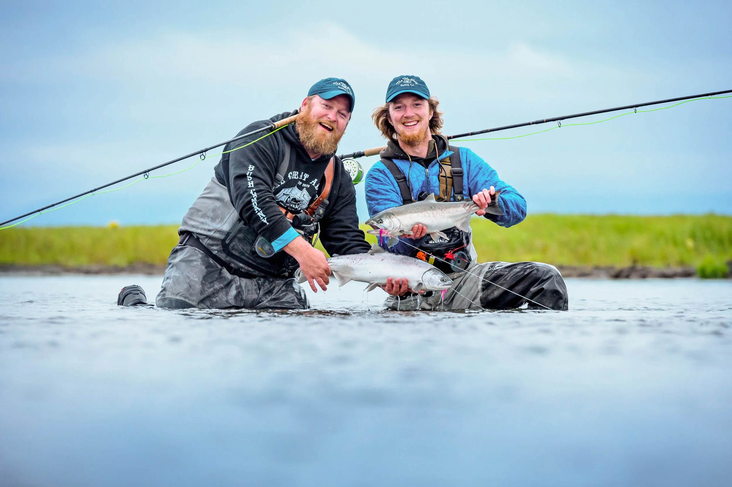 Two men kneeling in shallow water, holding caught fish, with fishing rods on their backs, outdoors near a grassy area under a cloudy sky.