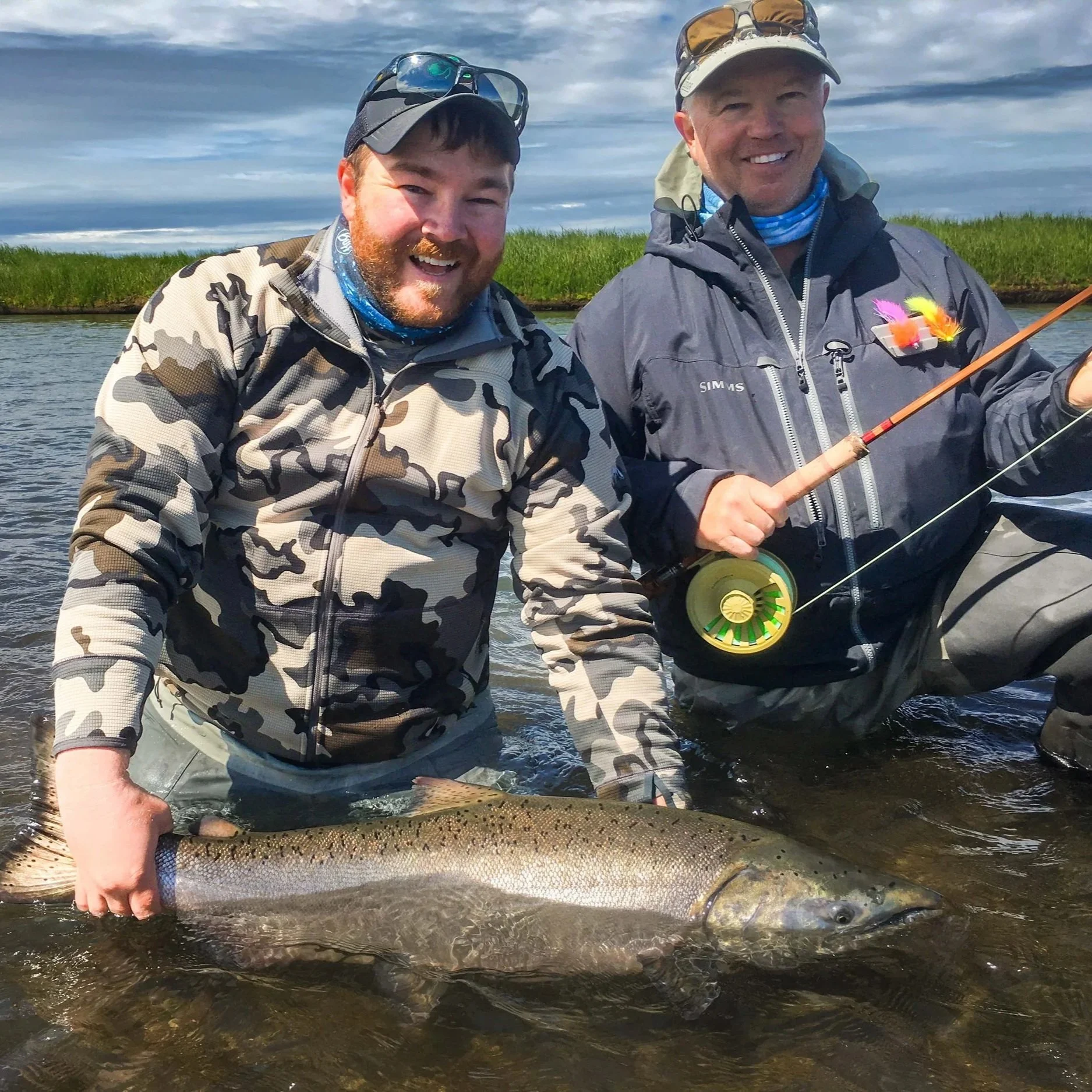 Two men in outdoor fishing gear holding a large fish in a river, with grassy banks and cloudy sky in the background.