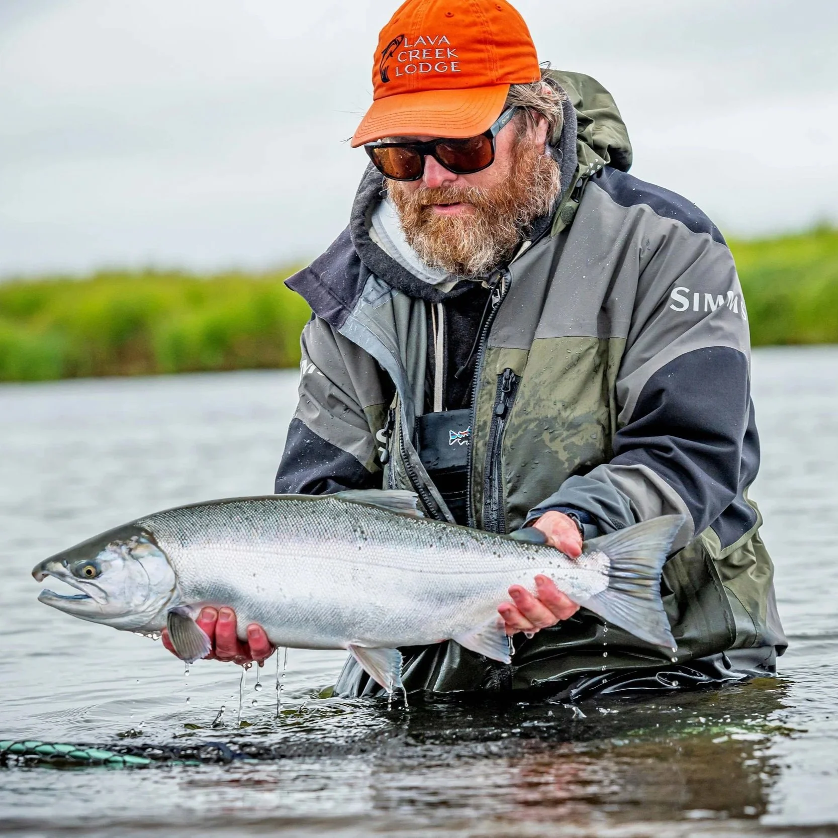 A man with a beard wearing sunglasses, an orange hat with 'Lava Creek Lodge' written on it, and outdoor gear, holding a large fish in a river.