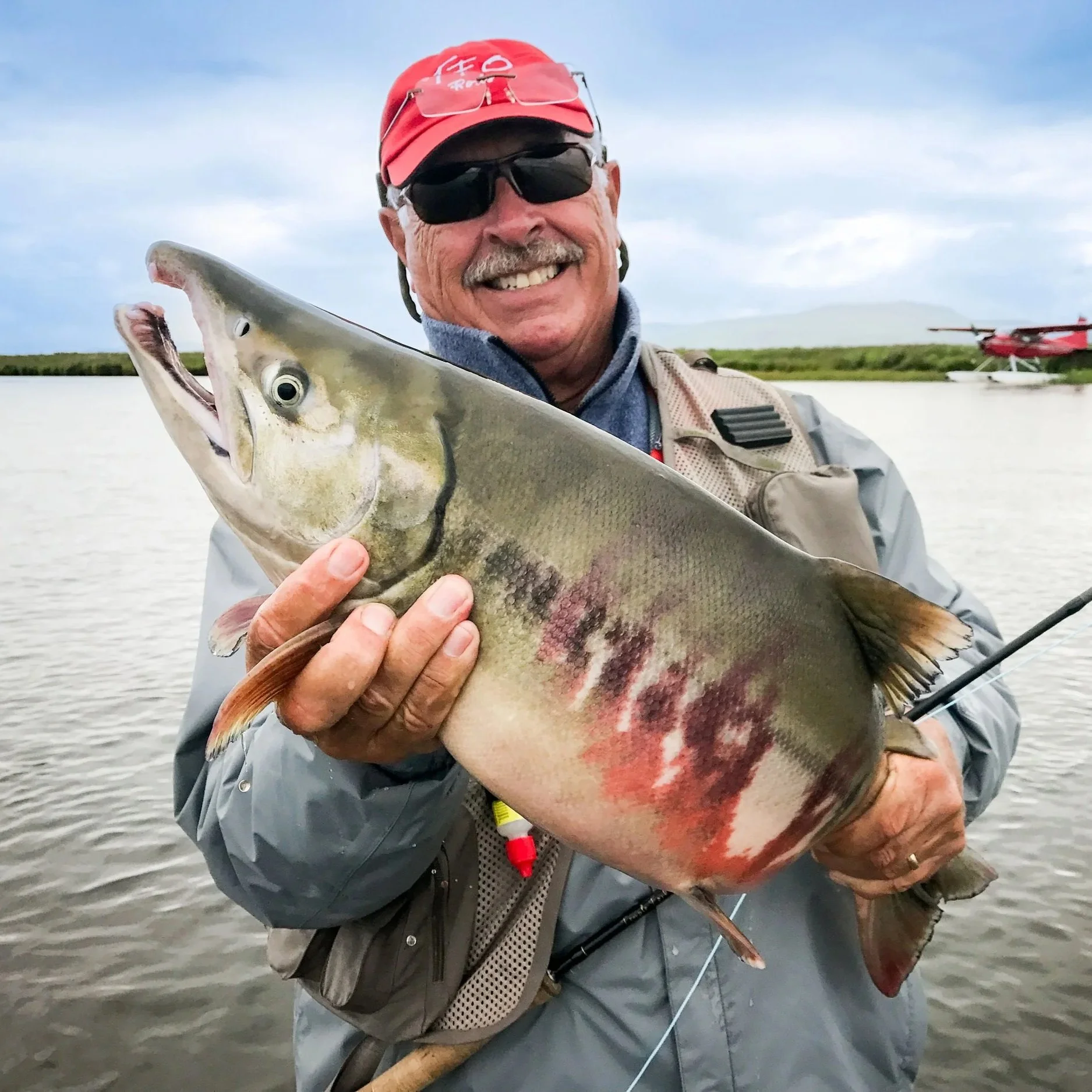 A man in sunglasses and a red cap holding a large fish in front of a body of water with an airplane in the background.
