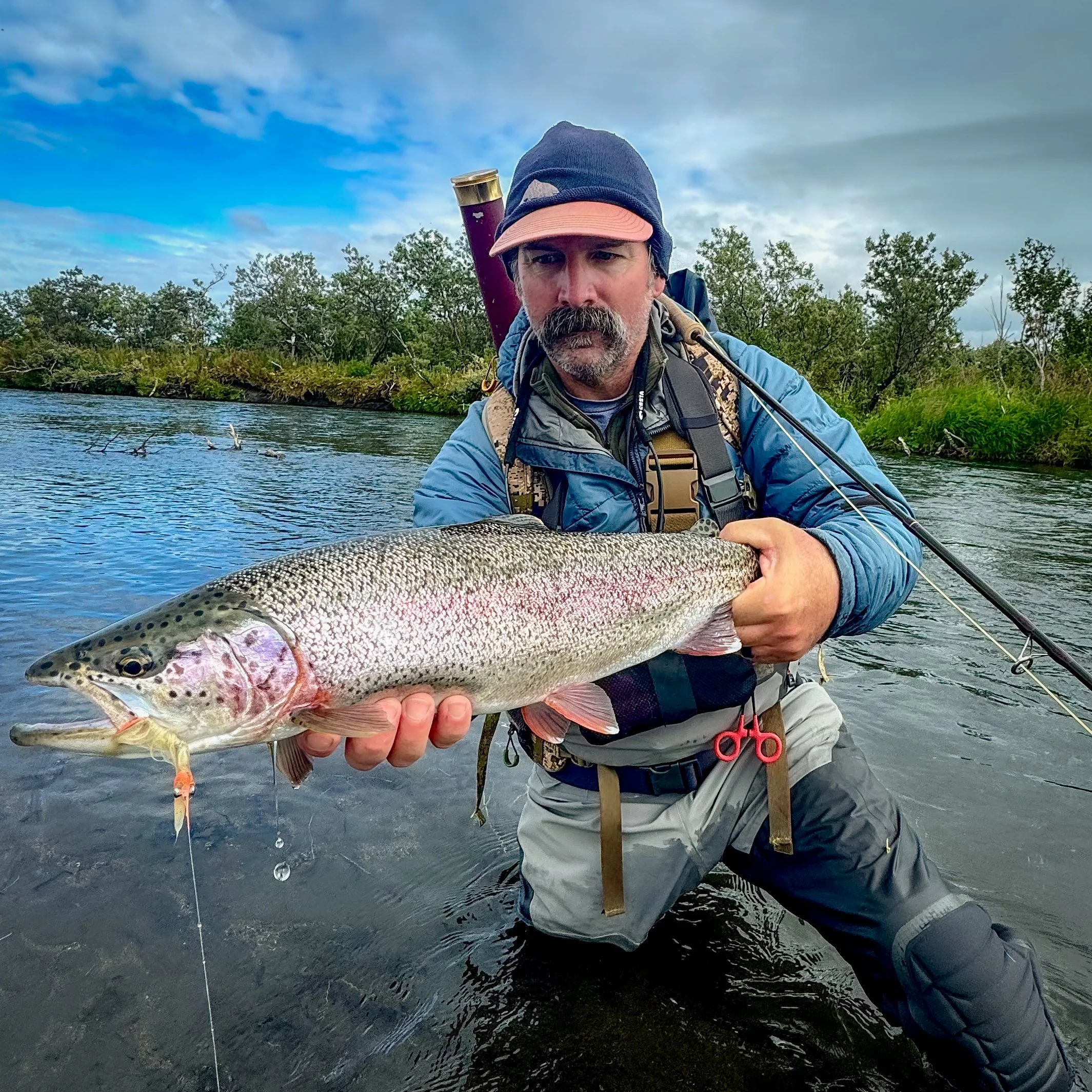 A man wearing outdoor gear, including a black beanie and a blue jacket, holding a large fish near a river with trees in the background.