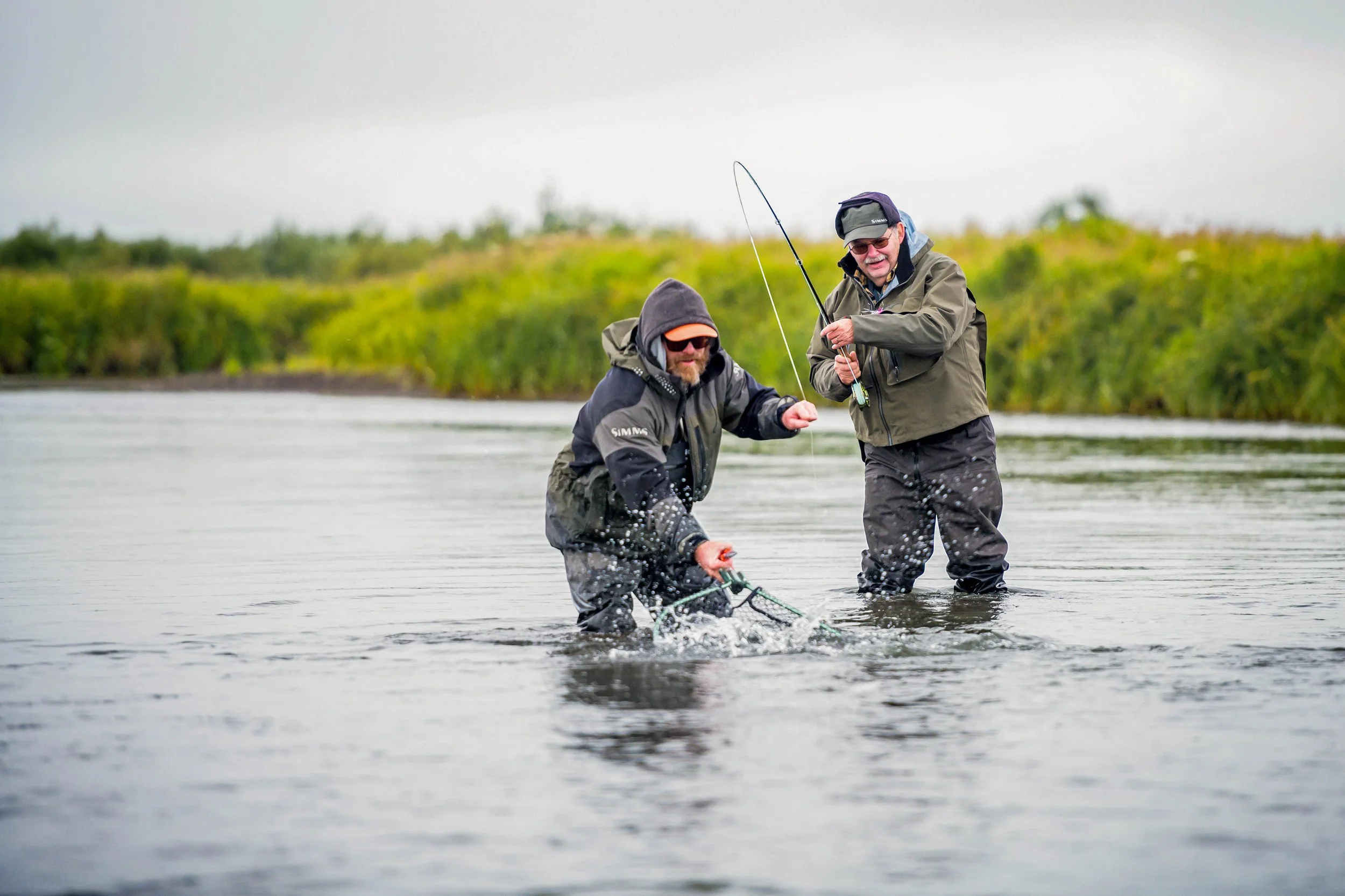 Two men wearing outdoor gear and waders standing in a river, fishing with rods, with green vegetation and a cloudy sky in the background.