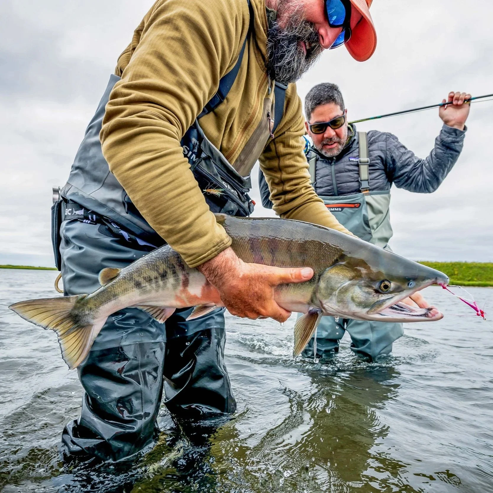 Two men in waders and sunglasses fishing in a river. One man is holding a large fish, and the other is holding a fishing rod.