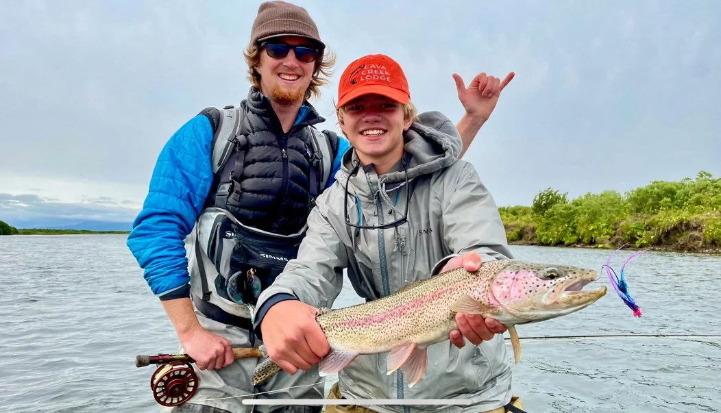 Two smiling young men on a riverbank, one holding a large fish with a fishing lure in its mouth, with trees and cloudy sky in the background.
