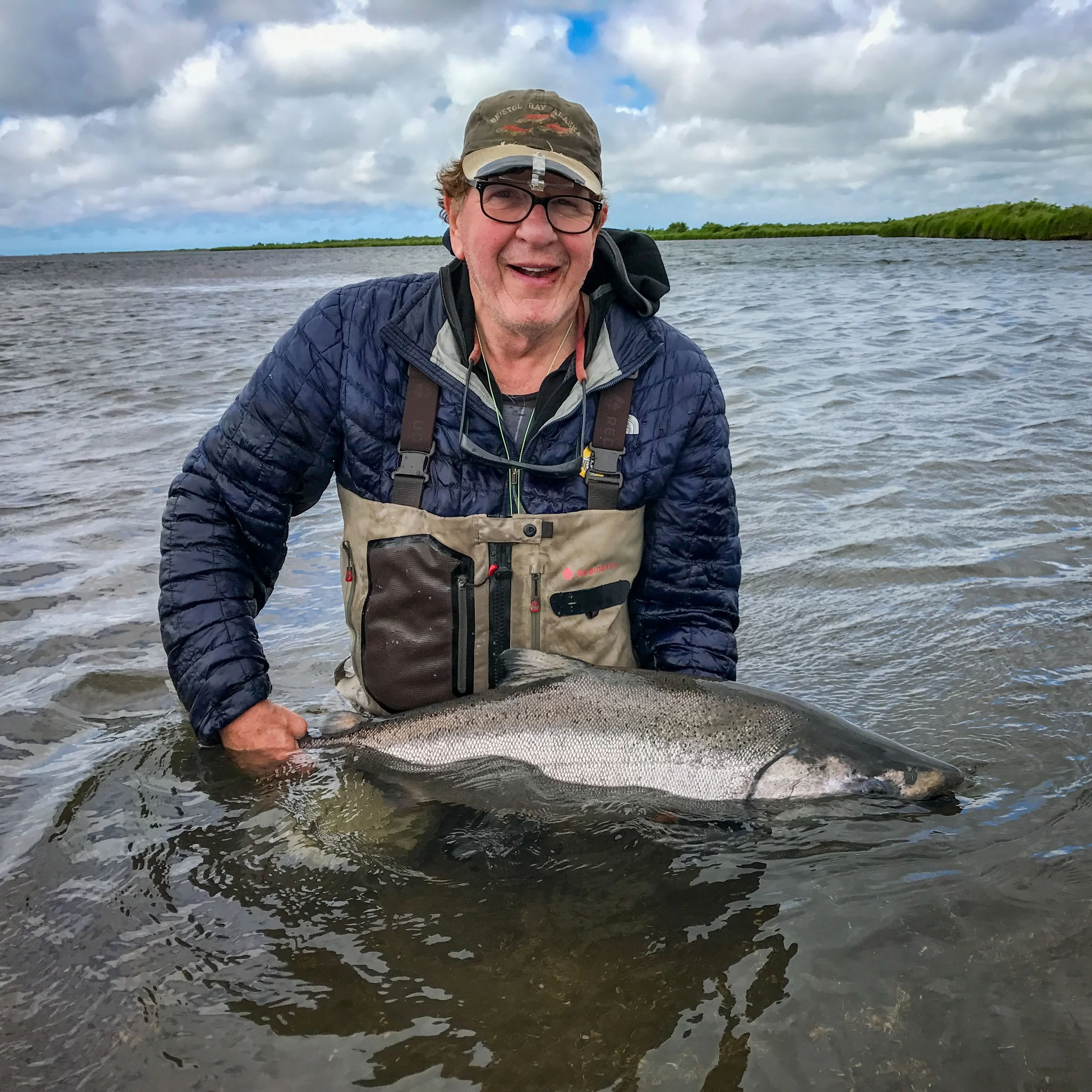 A man wearing a baseball cap, glasses, a blue quilted jacket, and a fishing vest is smiling and holding a large fish near the water, with a cloudy sky and green shoreline in the background.