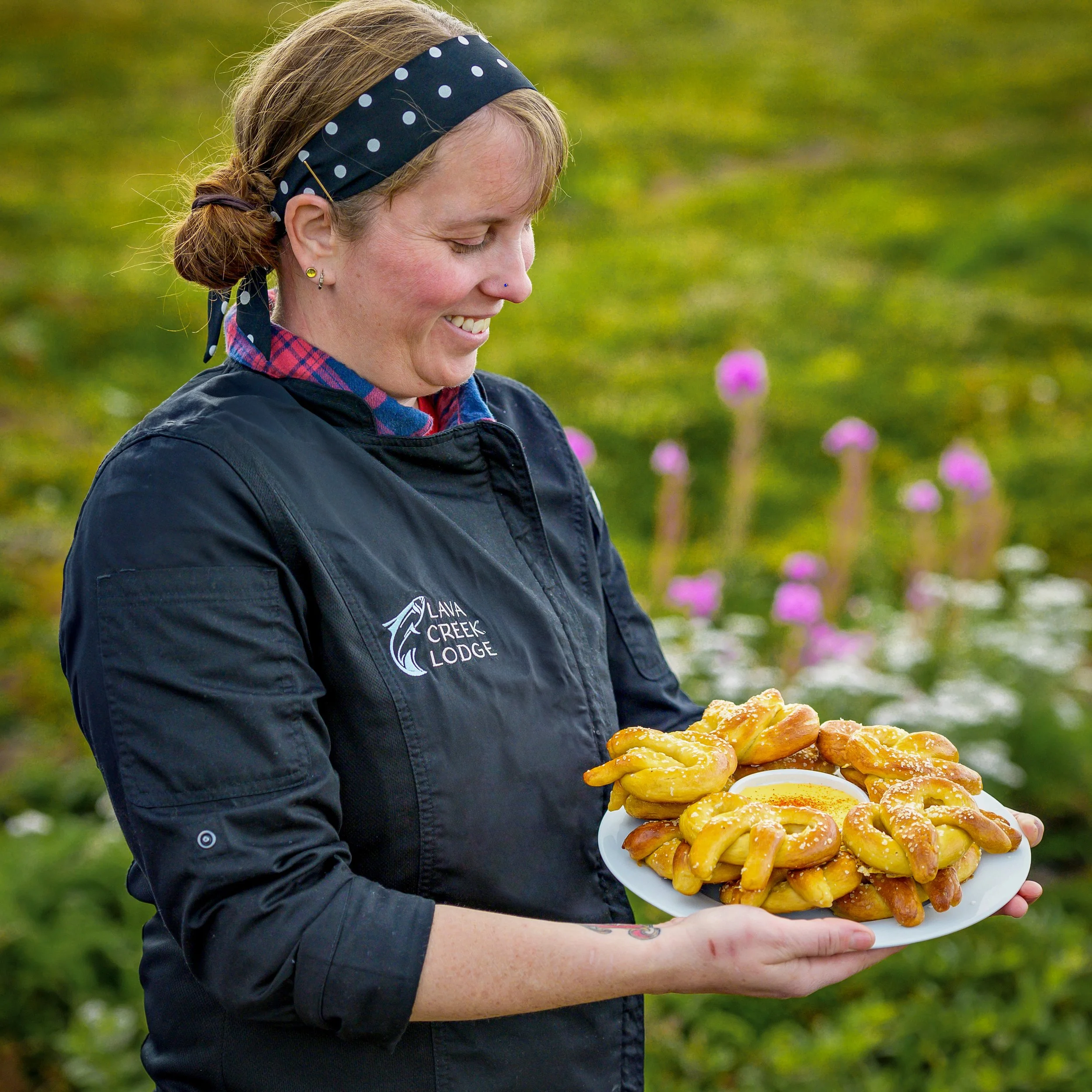 A woman with a polka dot headband and earrings is smiling while holding a large plate of funnel cakes topped with powdered sugar, outdoors in a grassy area with purple flowers in the background.
