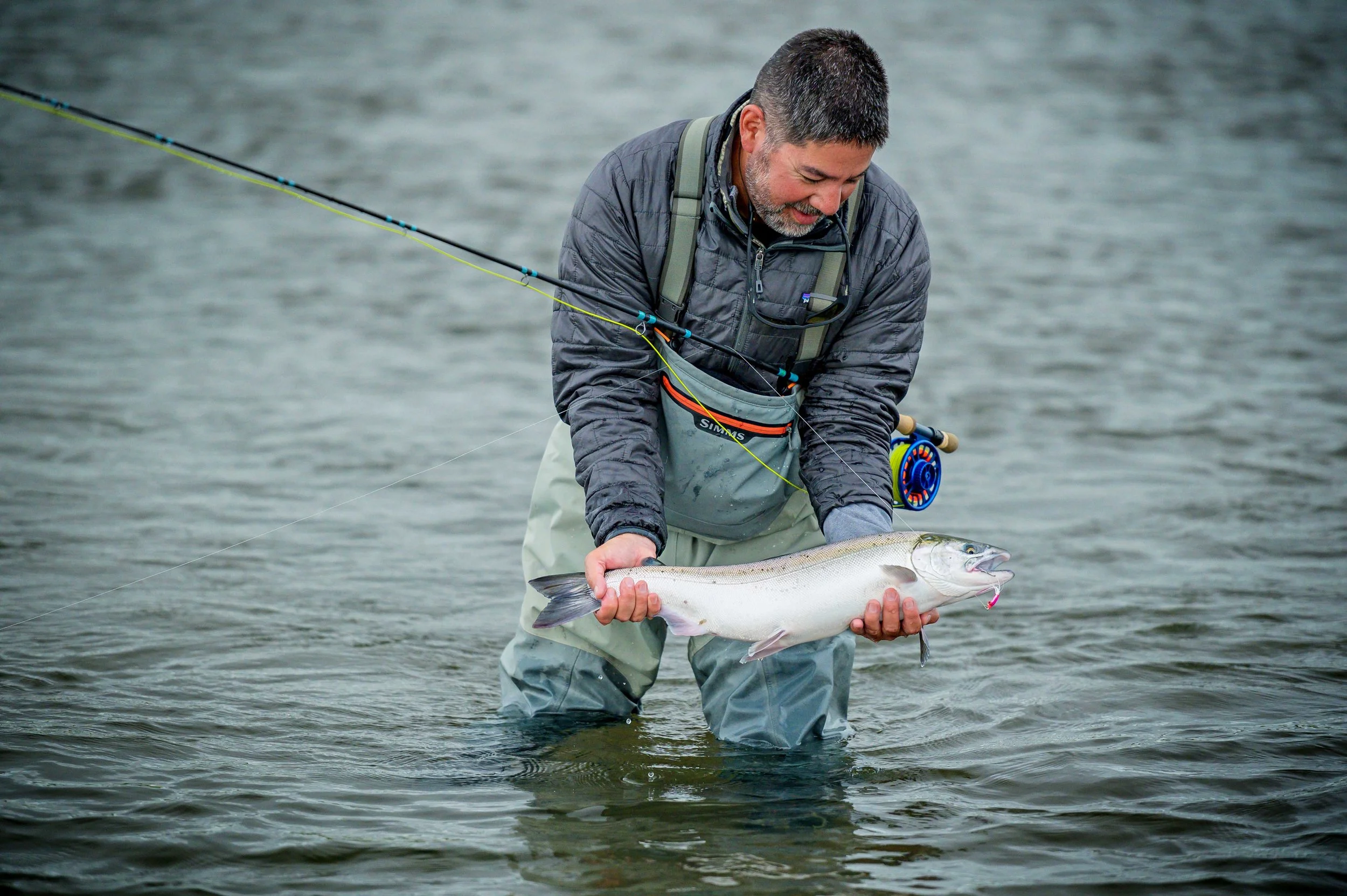 A man wearing outdoor fishing gear stands in shallow water, holding a large fish with both hands. He appears to be examining the fish after catching it.