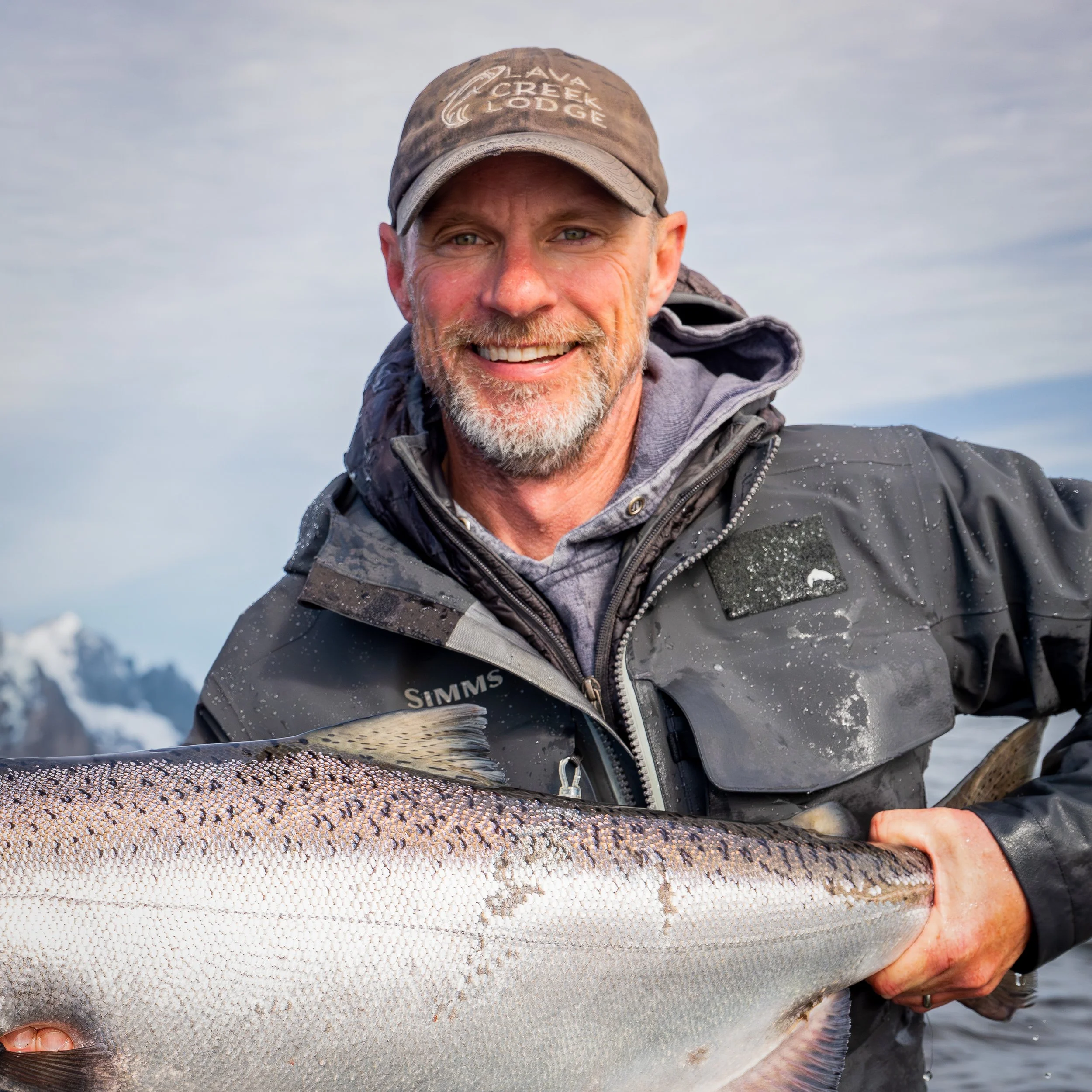 A smiling man wearing a brown cap and outdoor gear holding a large fish with silver scales and reddish fins.