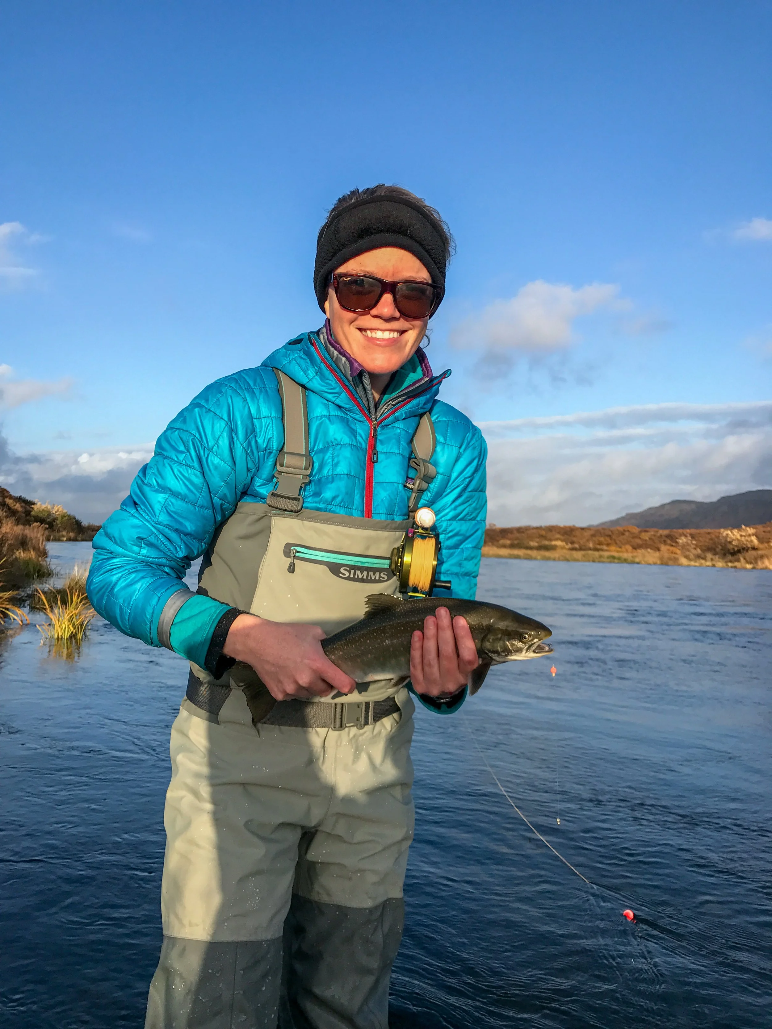 A person dressed in outdoor fishing gear, holding a fish, standing in a body of water with mountains and a clear sky in the background.