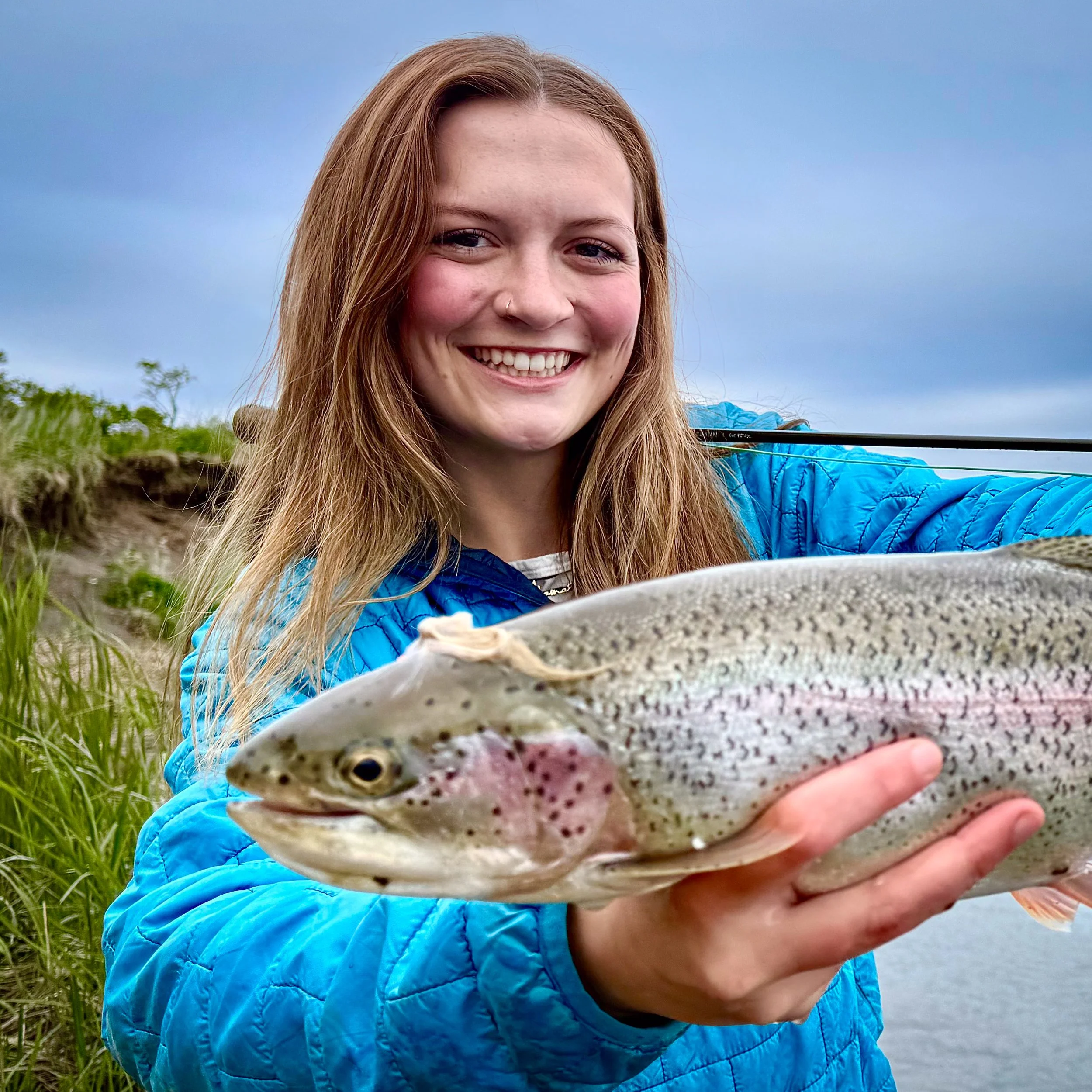A young woman with long red hair smiling and holding a freshly caught rainbow trout fish outdoors, with a cloudy sky and greenery in the background.