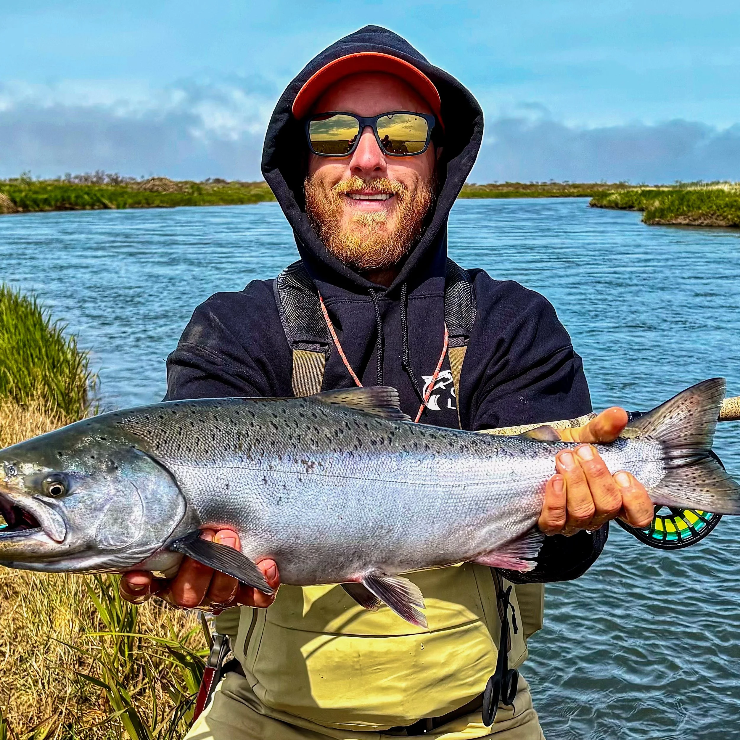 A man with a beard wearing sunglasses, a black hooded jacket, and a red cap holding a large fish near water and greenery on a sunny day.