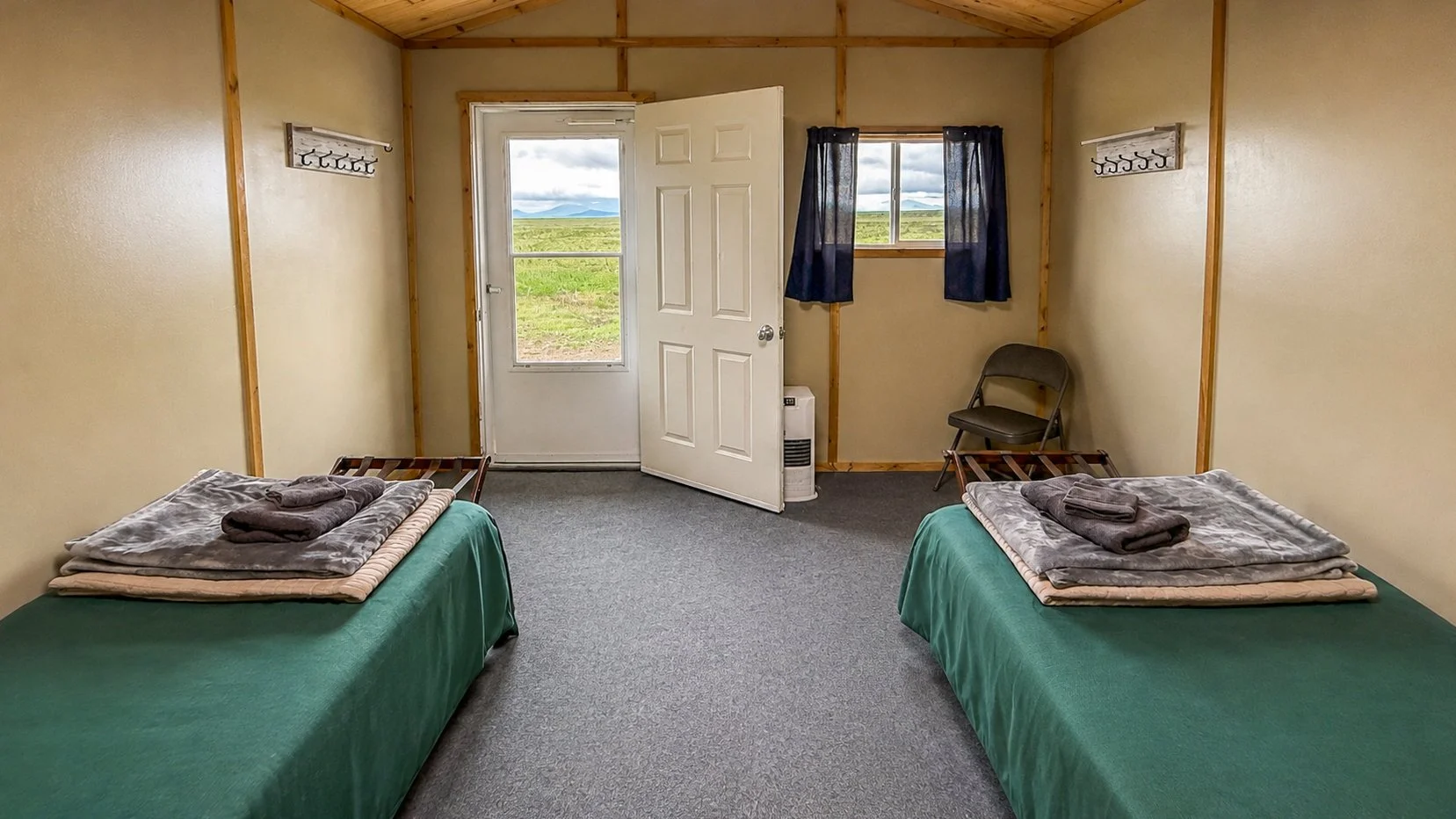A simple motel room with two beds, each with dark green bedspreads and folded towels on top. The room has beige walls with wood trim, a small window with dark curtains, and a door leading outside showing a grassy landscape and mountains in the distance. There's a single chair, a wall-mounted coat rack, and a small space heater near the door.