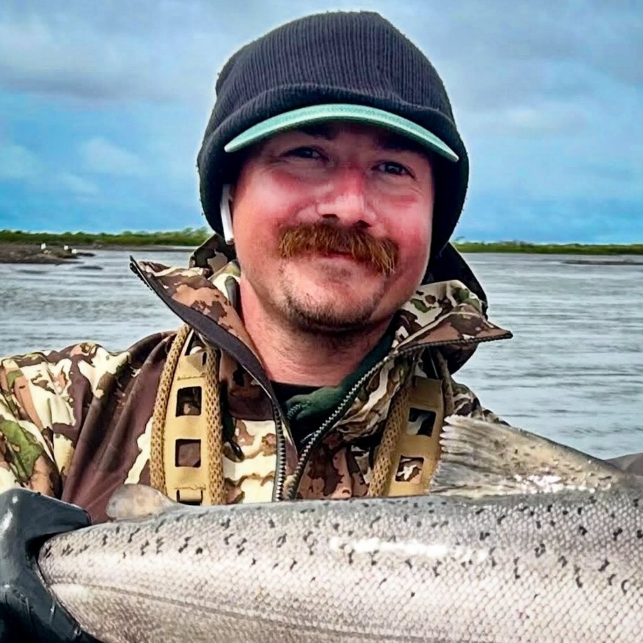 Man outdoors in camo jacket holding a large fish, with water and cloudy sky in the background.