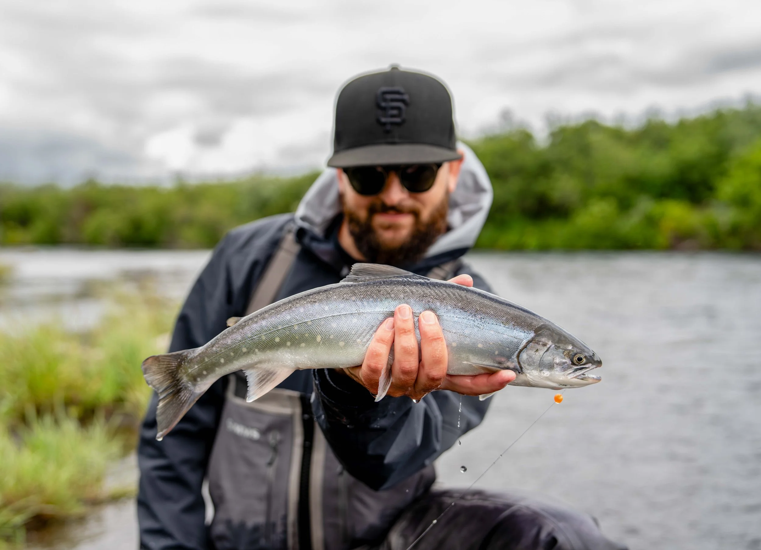 Man holding a large fish outdoors near water with green trees and cloudy sky in the background.