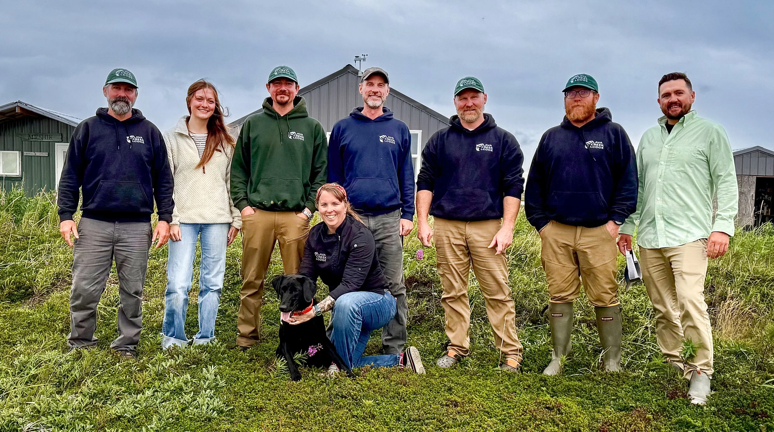 Group of nine people and a dog outdoors in front of a metal building under cloudy sky. Most wear hoodies or jackets with 'Lava Creek Lodge' logo. One woman kneels with a black Labrador Retriever.