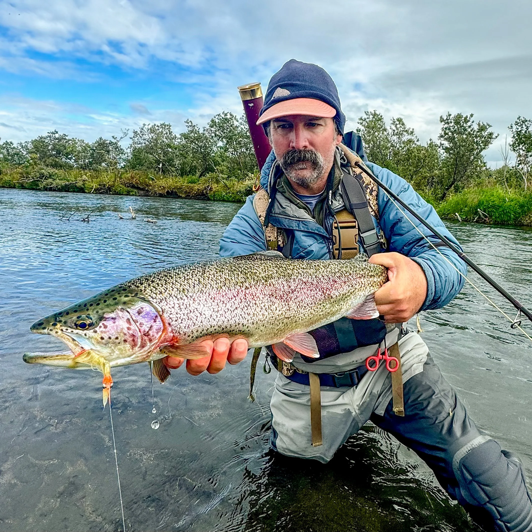 A man with a gray beard and mustache wearing a blue jacket, dark beanie, and tan cap, holding a large fish with silver, green, and black scales, near a river with trees and cloudy sky in the background.