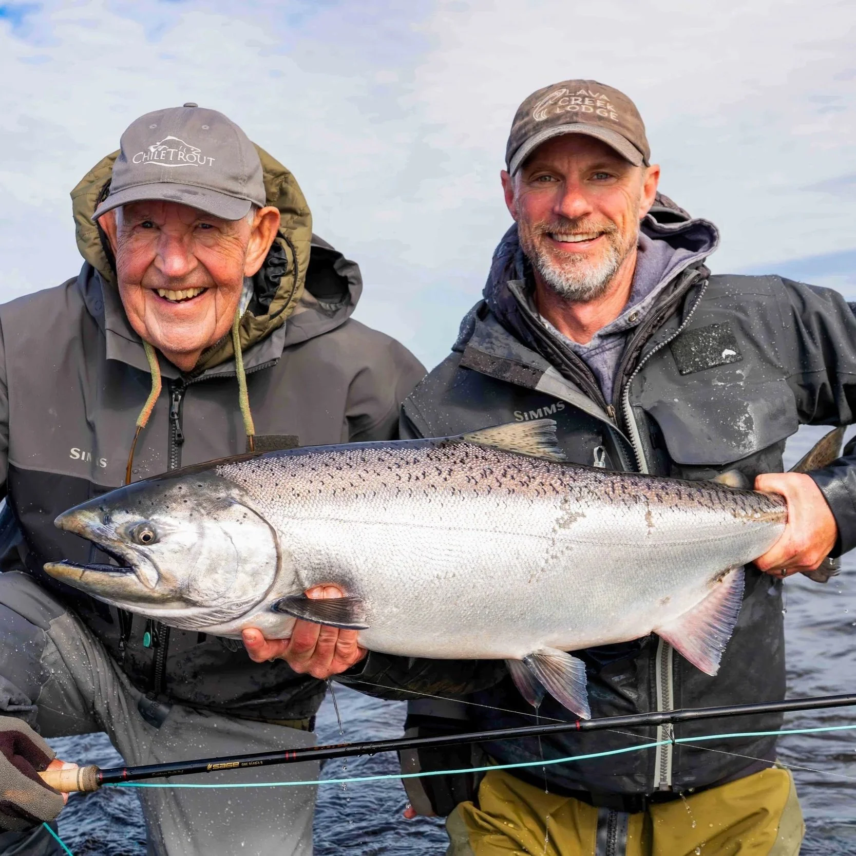 Two men holding a large fish, boat fishing gear visible, outdoors with water and cloudy sky on the Alaska Peninsula at Lava Creek Lodge.