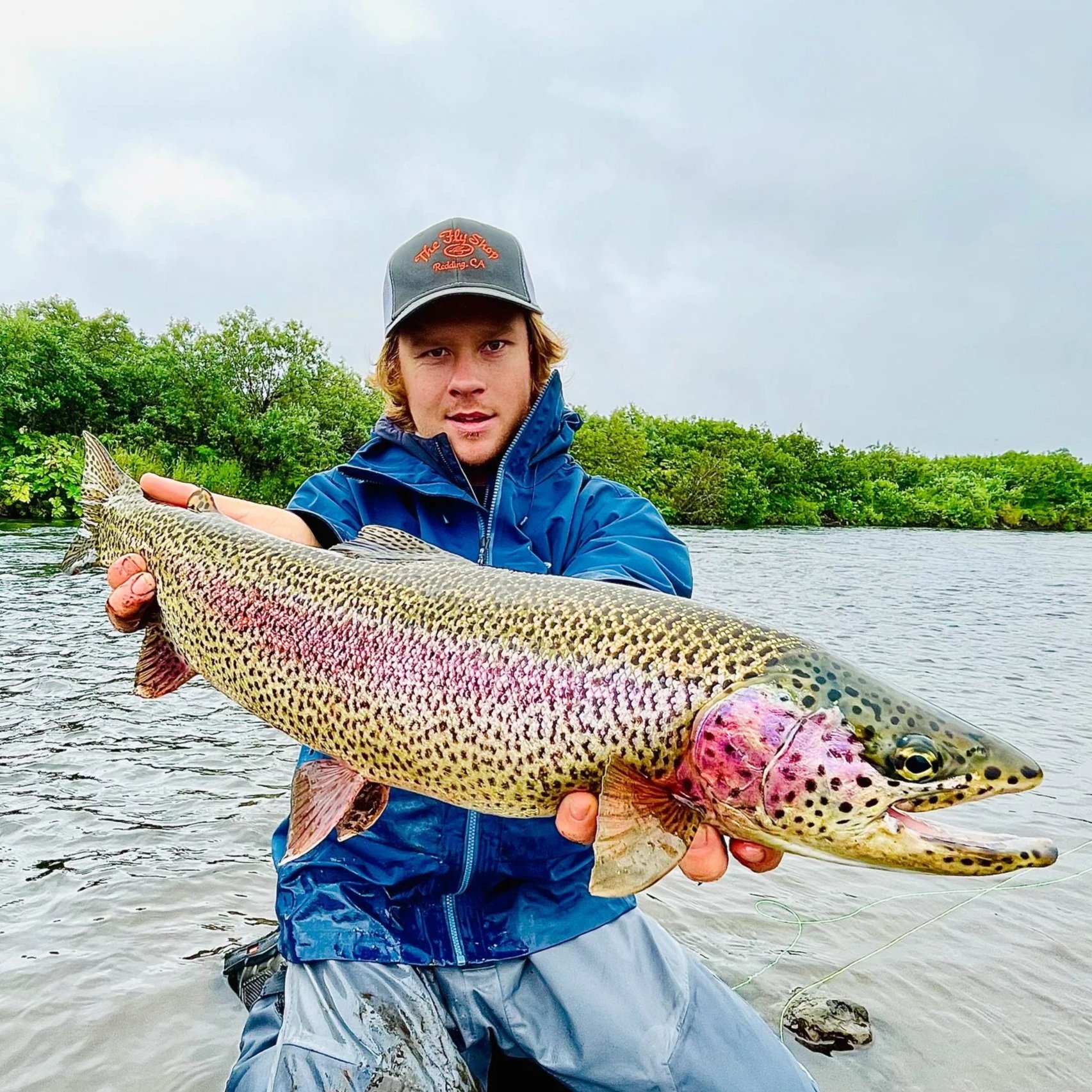 A man wearing a baseball cap and a blue waterproof jacket holding a large trout fish near a river with green trees in the background.