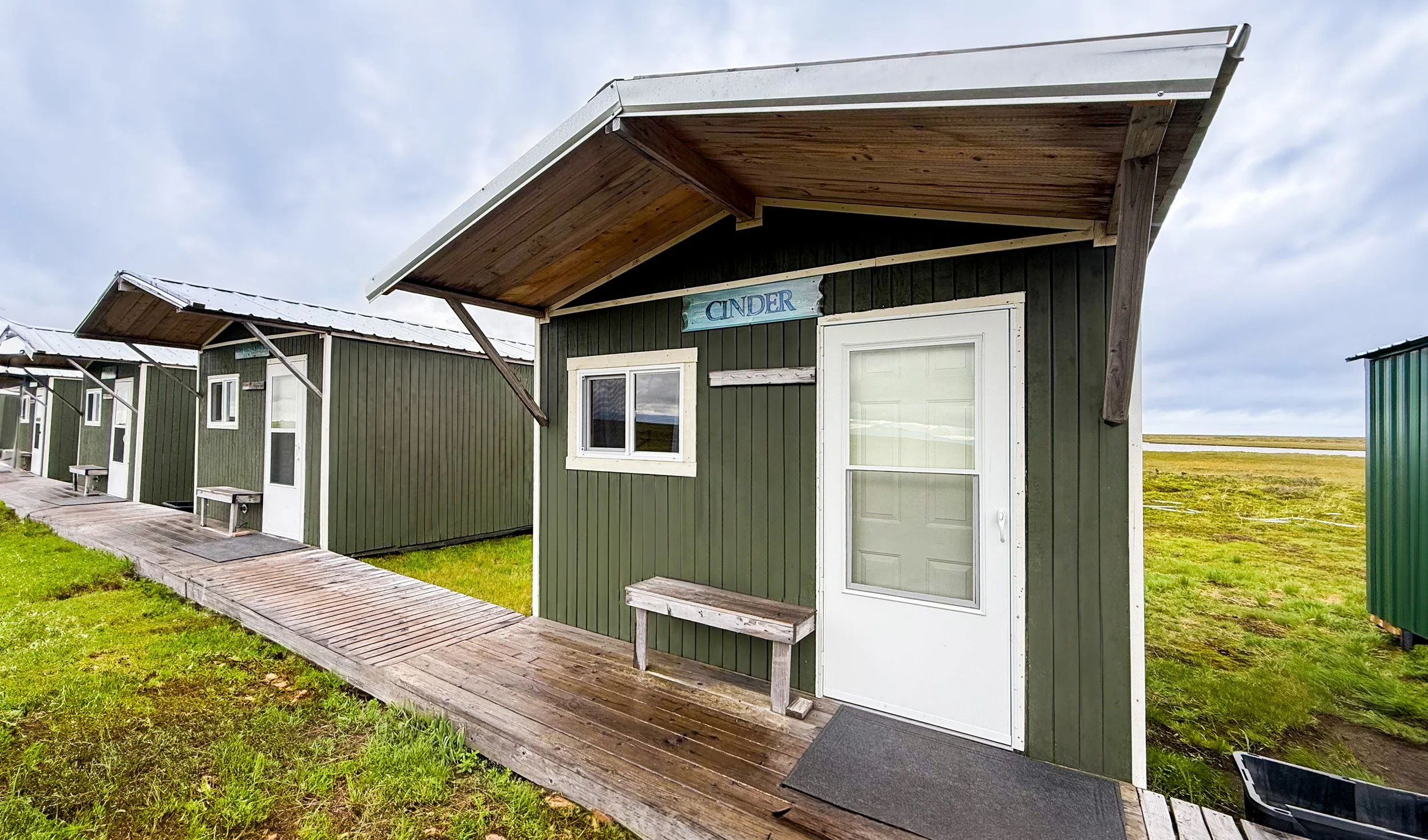 A tiny house with dark green siding, a white door, and a small window, sitting on a wooden porch with a bench, under a cloudy sky.