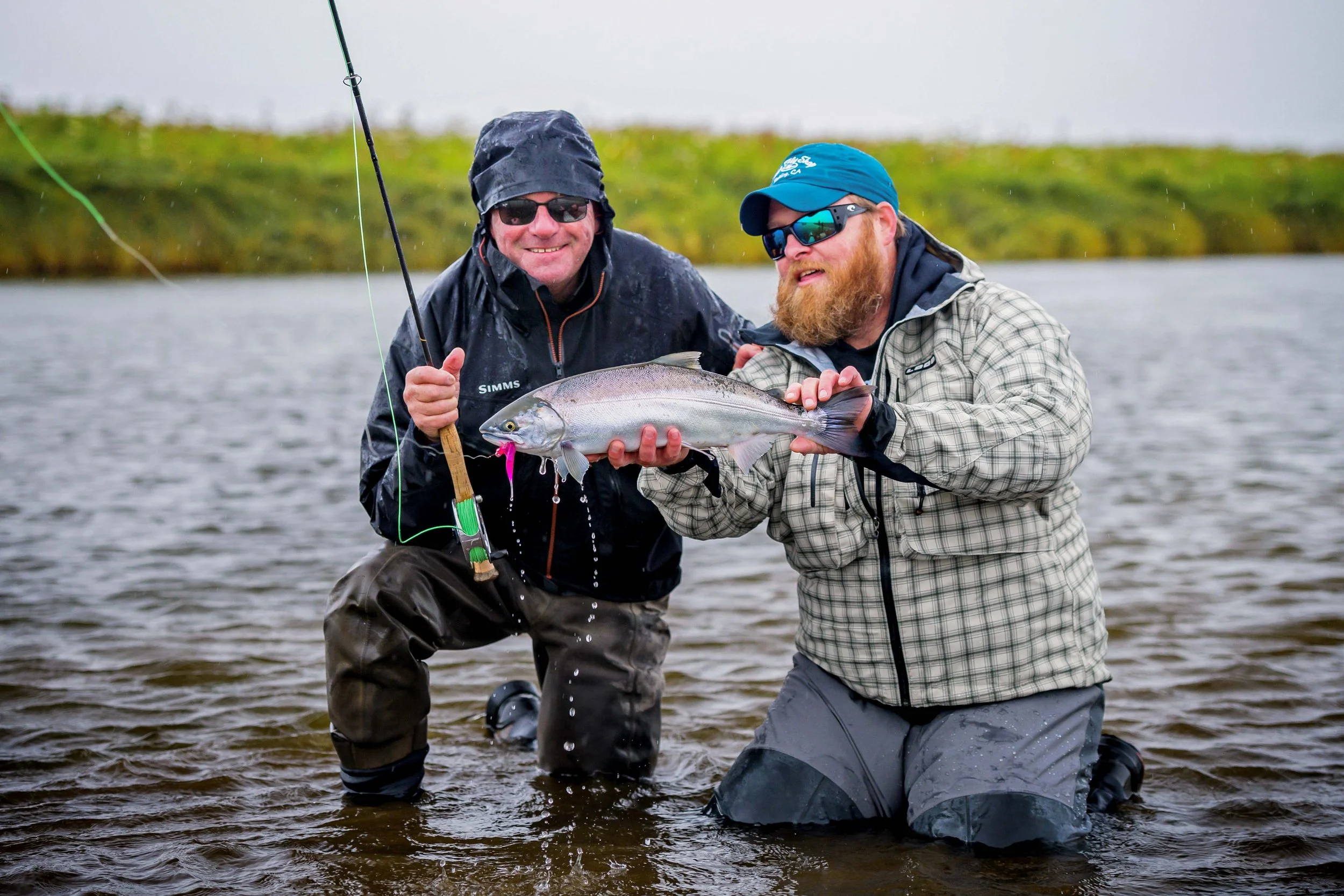 Two men in waterproof clothing and sunglasses are kneeling in a river, holding a large fish they caught together during a fishing trip.