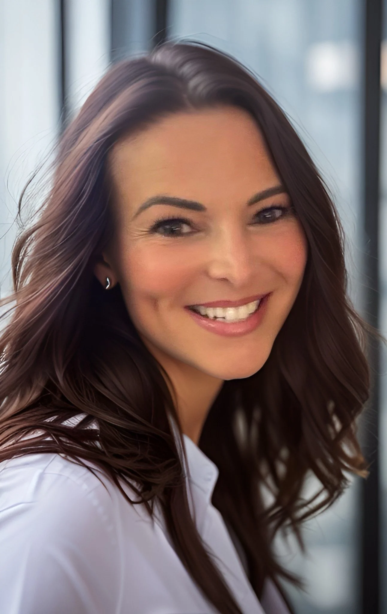 Close-up of a woman smiling with long, wavy brown hair, wearing a white shirt.