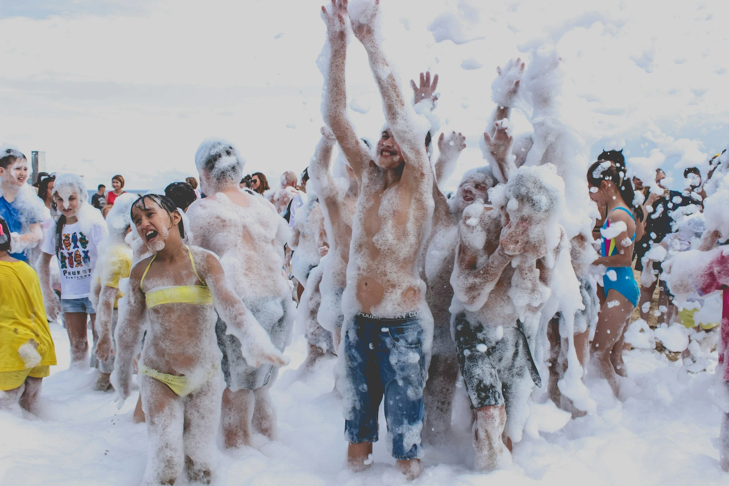 Group of children and a man enjoying a foam party at the beach, playing in the foam and smiling.