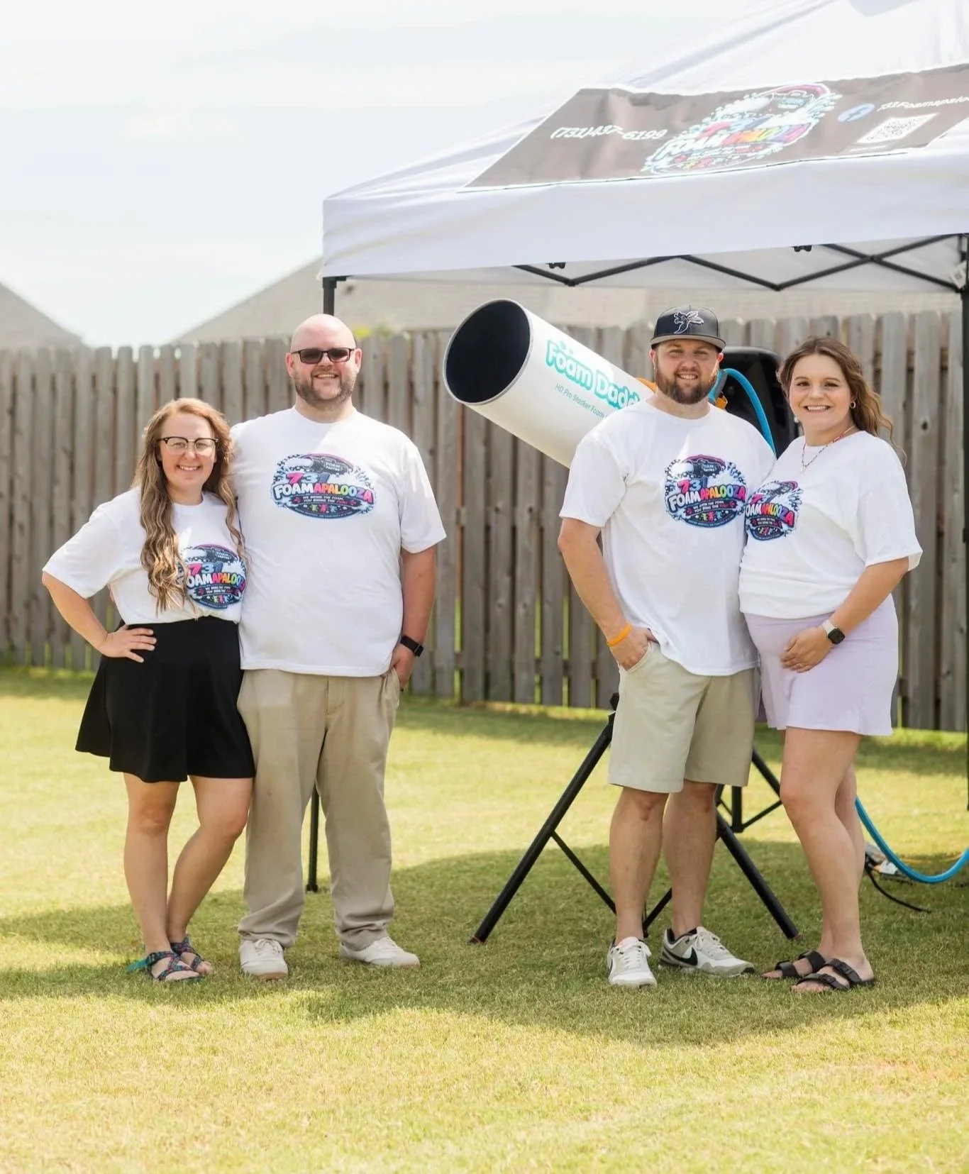 Four people standing on a grassy backyard, smiling for a photo. They are wearing white T-shirts with a colorful logo that reads 'Foamapalooza'. There is a wooden fence and a white tent behind them, with a large foam cannon attached to a tripod, suggesting a foam or bubble event.