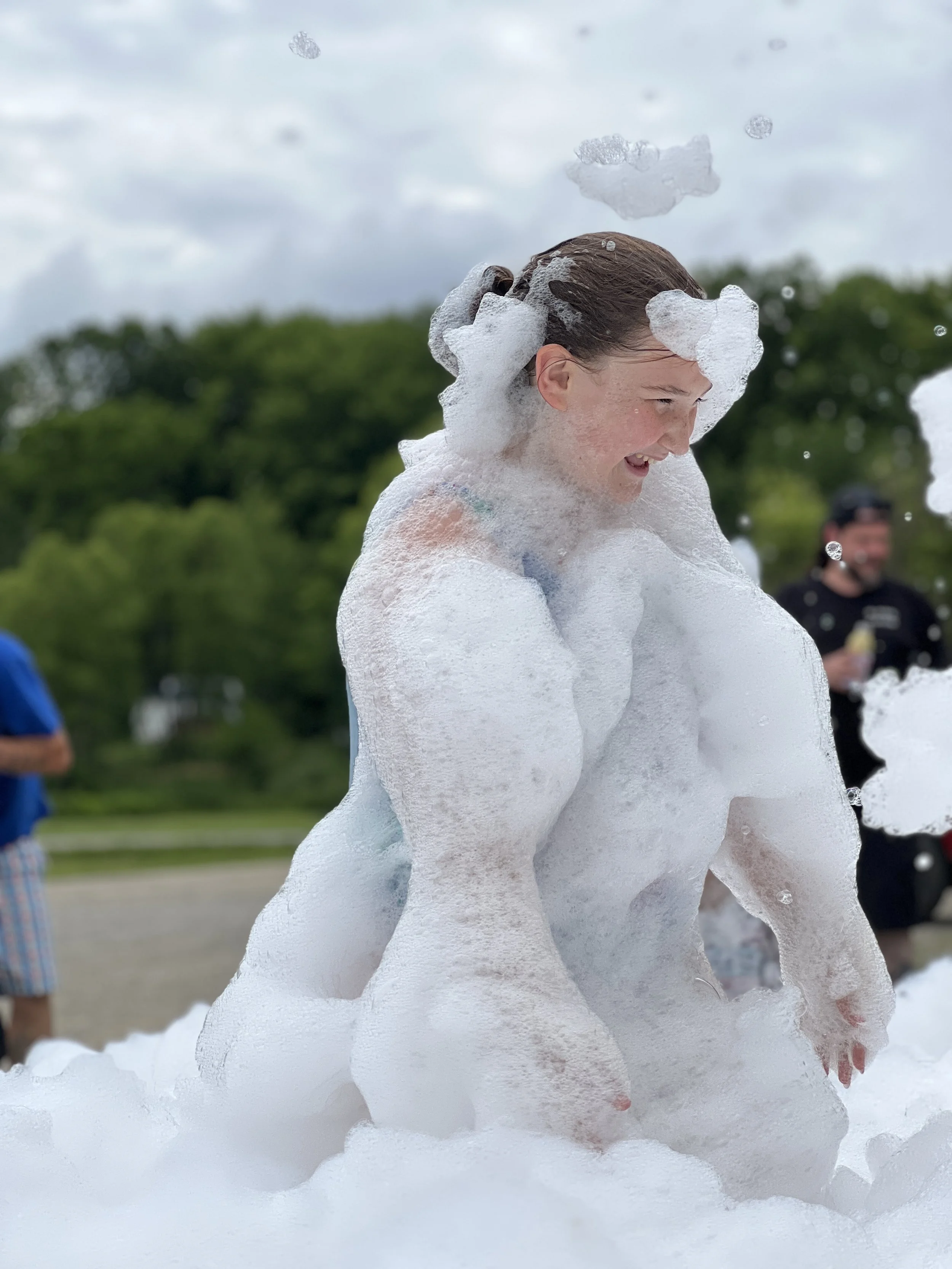 Person covered in white foam or bubbles in an outdoor setting with trees and cloudy sky in background.