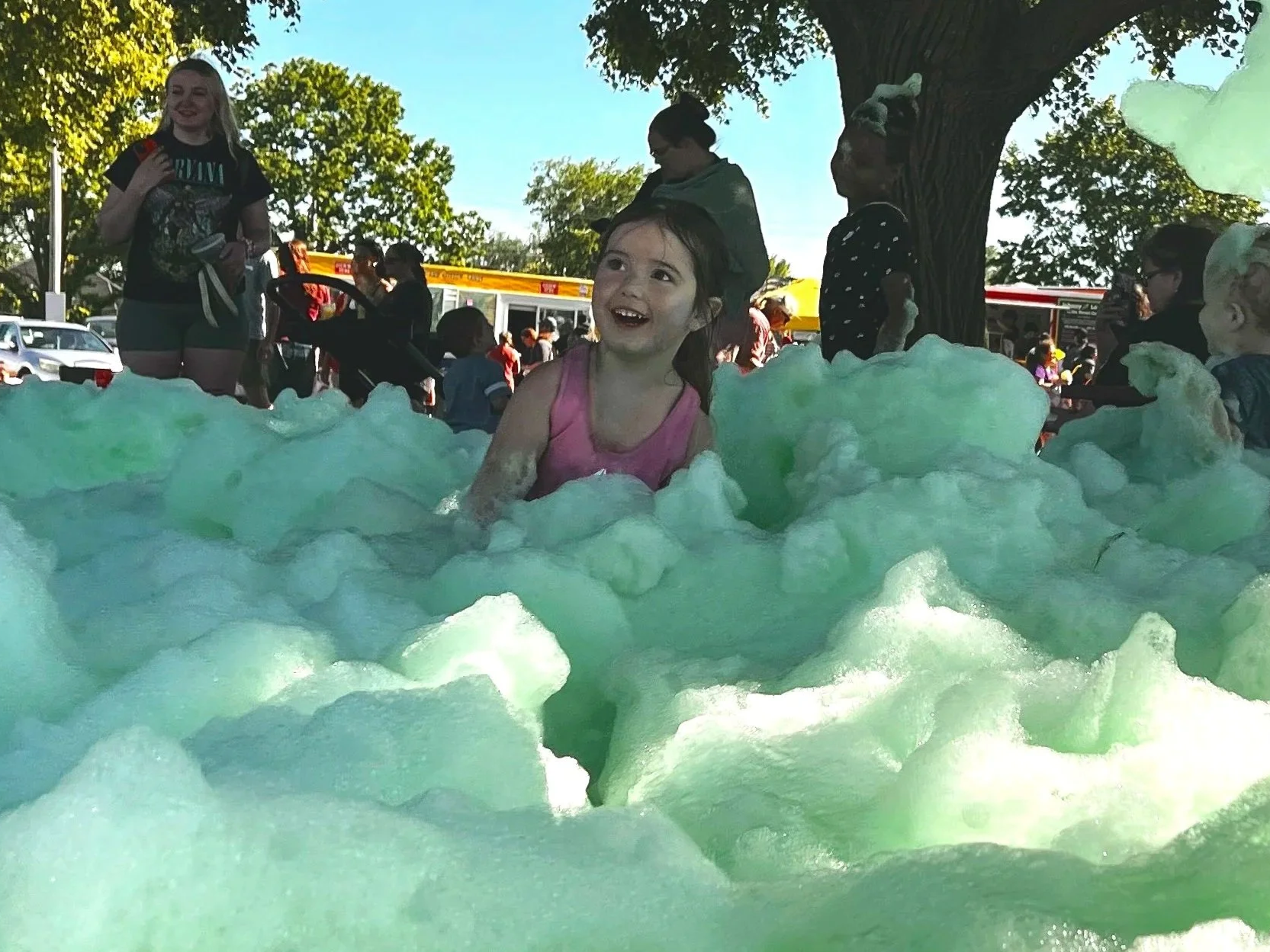 A young girl smiling and playing in a foam-filled area outdoors during daytime, with other people and trees in the background.