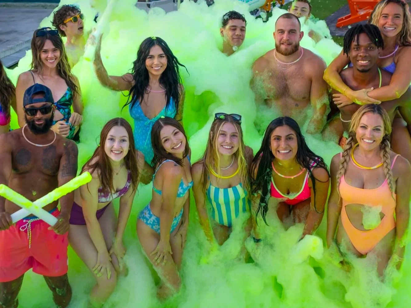 Group of young people in a foam party, smiling in swimsuits, surrounded by green foam.