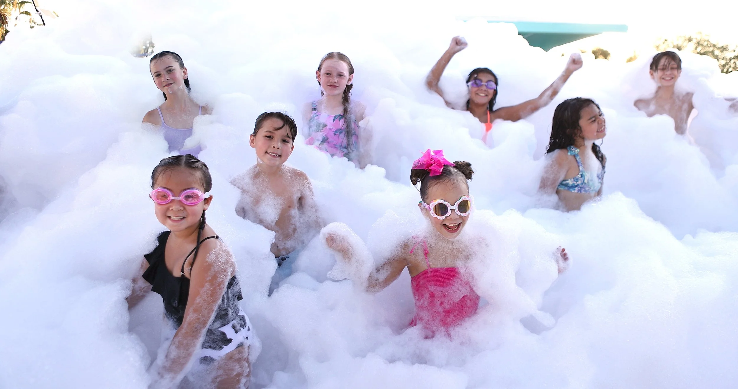 Children playing in a foam party, smiling and wearing swimsuits and goggles.