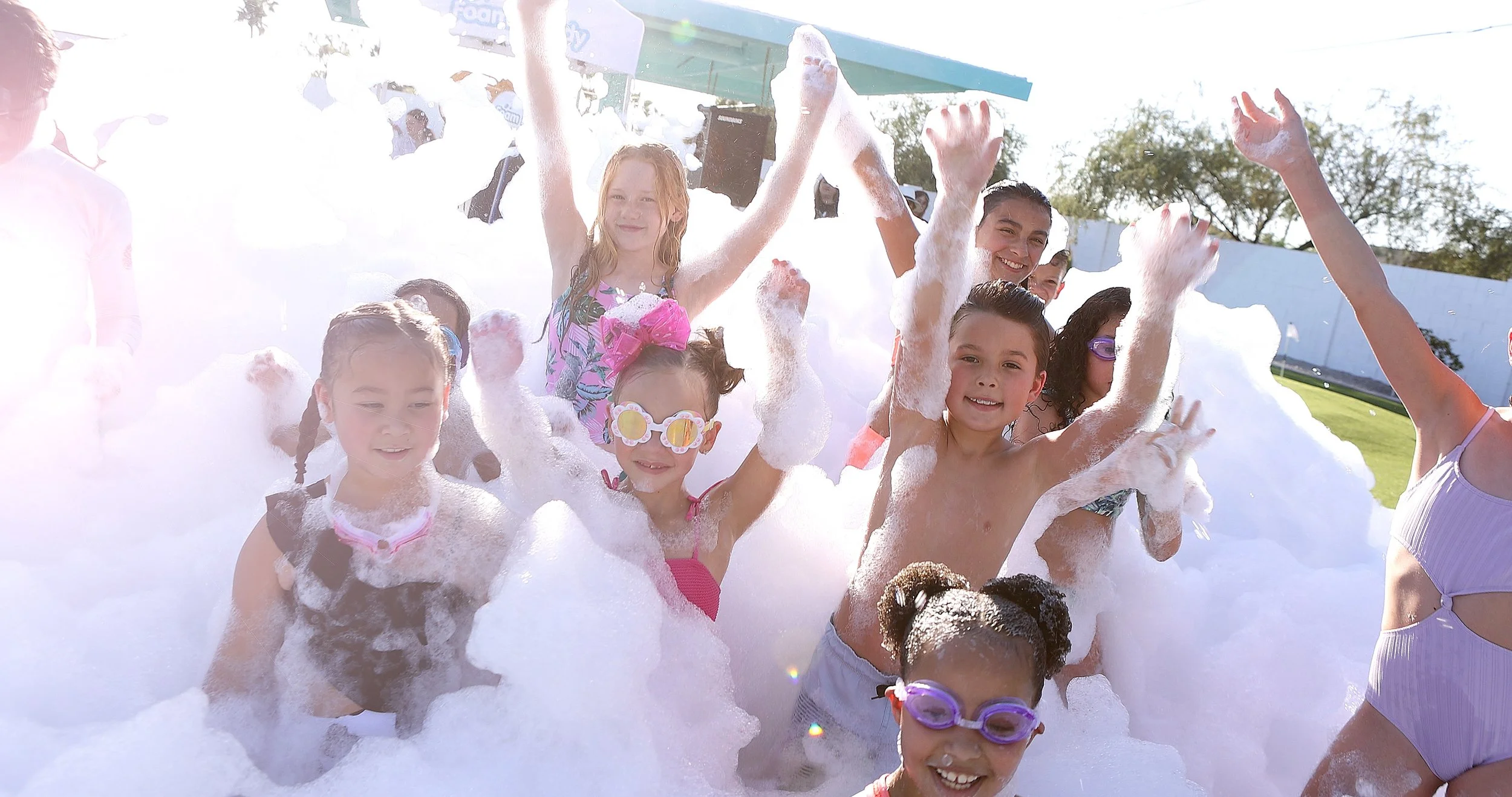 Group of children and a young woman having fun in a foam party outdoors on a sunny day.