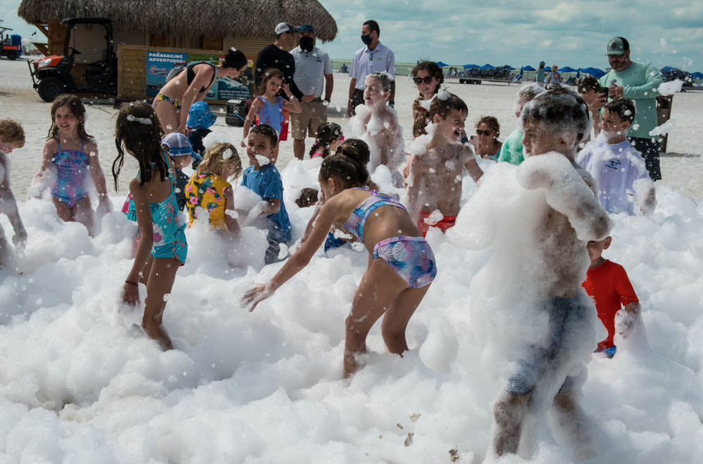 Children and adults playing in foam on a beach with blue sky, some people in swimsuits, and beach tents in the background.