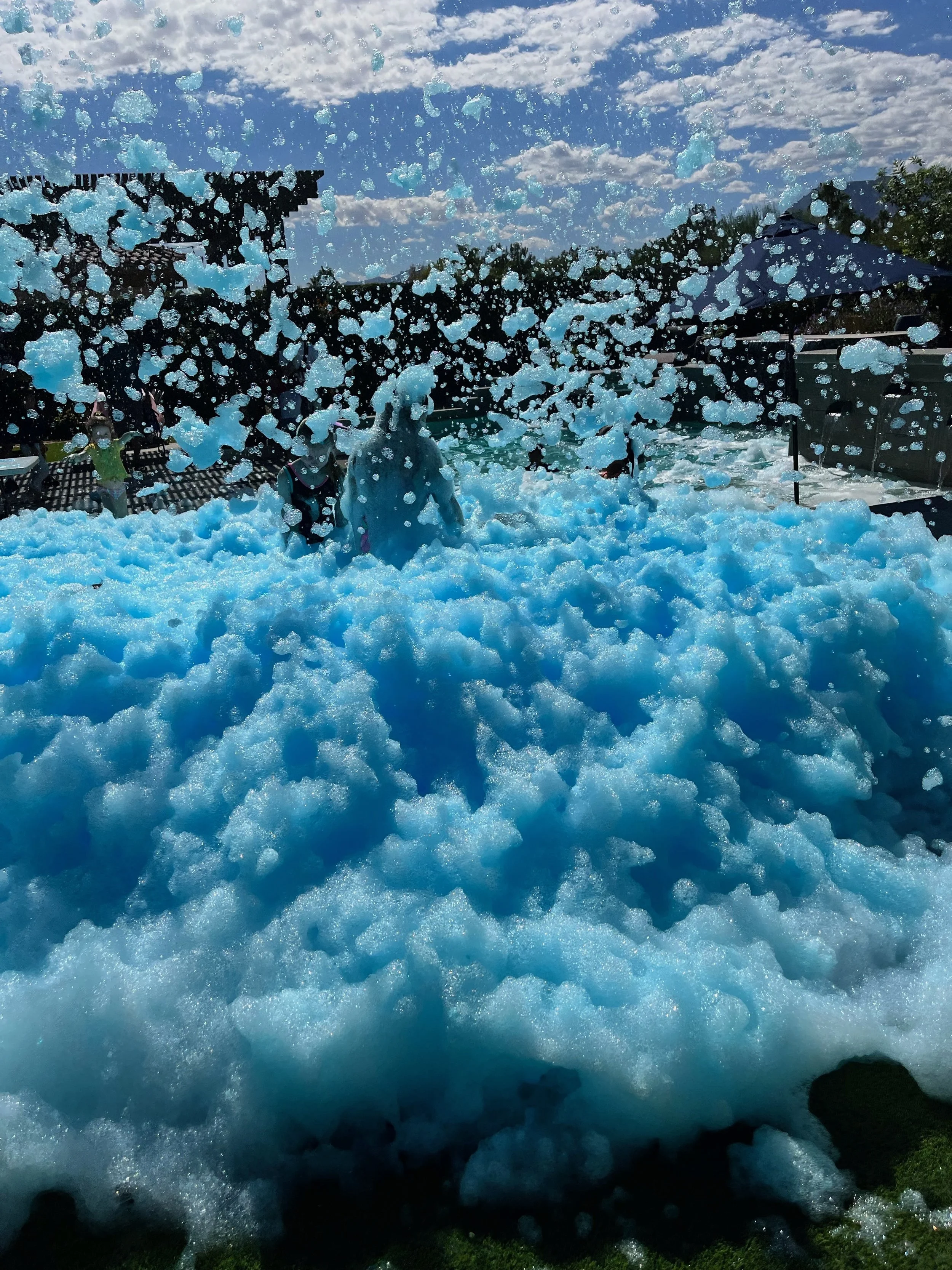 Foamy blue water waves crashing with a crowd of people in the background, under a sky with scattered clouds.