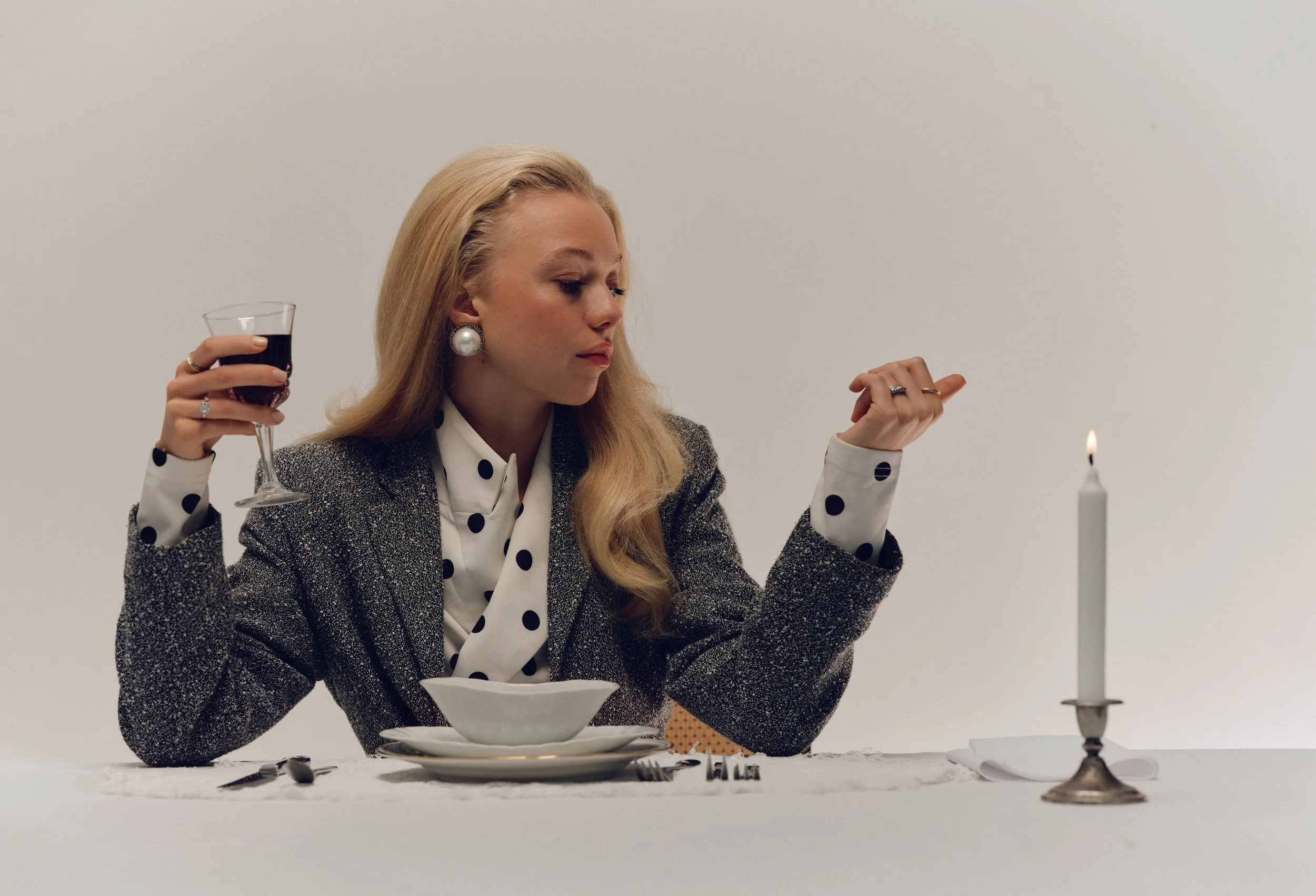 A woman with long blonde hair, large pearl earrings, and a polka dot shirt under a blazer, sits at a table set with a bowl, silverware, and a glass of red wine. She appears to be preparing to eat or in a contemplative mood, with a lit candle on the table against a plain white background.