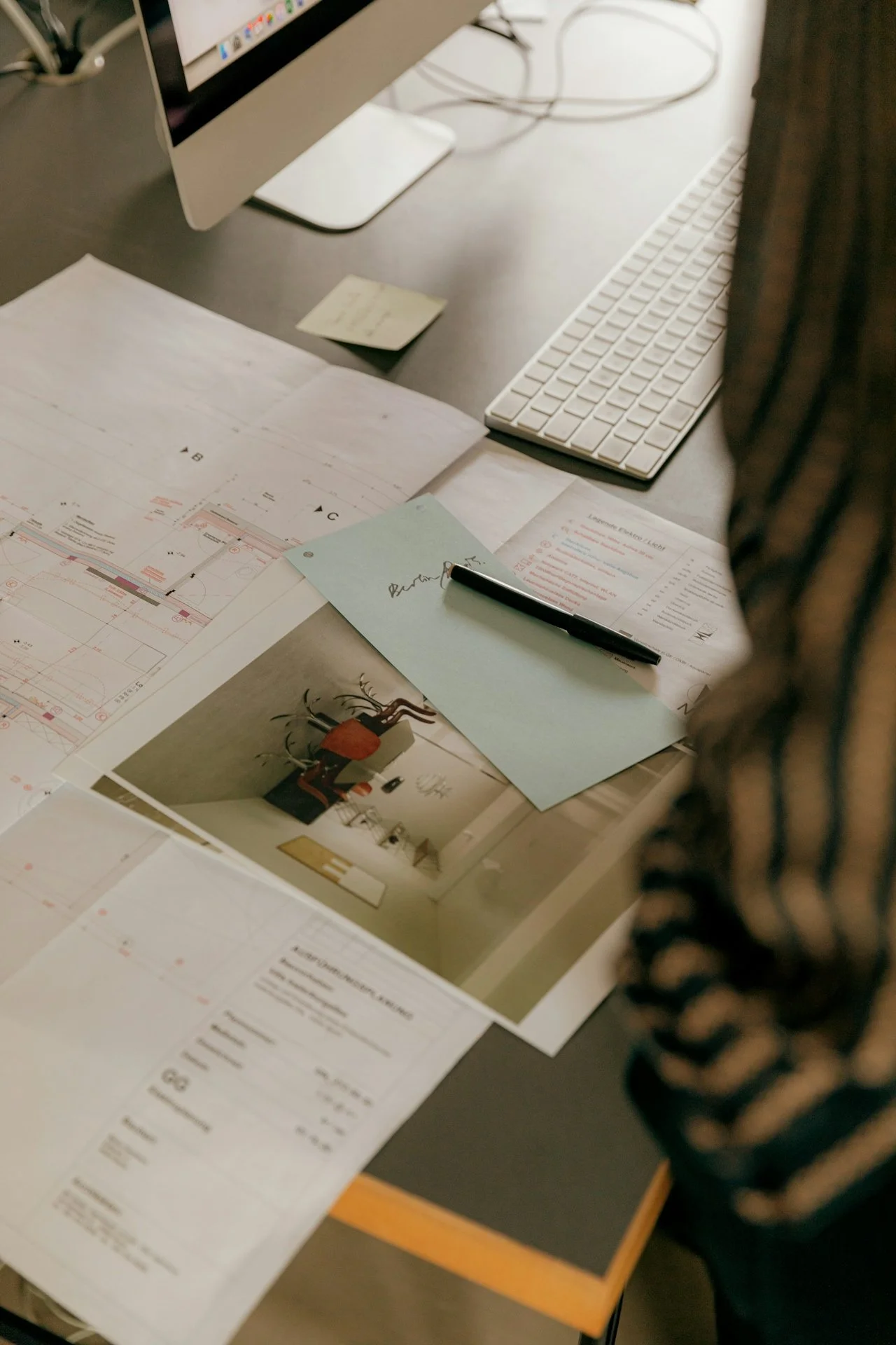 Desk with architectural plans, photographs, a computer, keyboard, notepad, and pen.