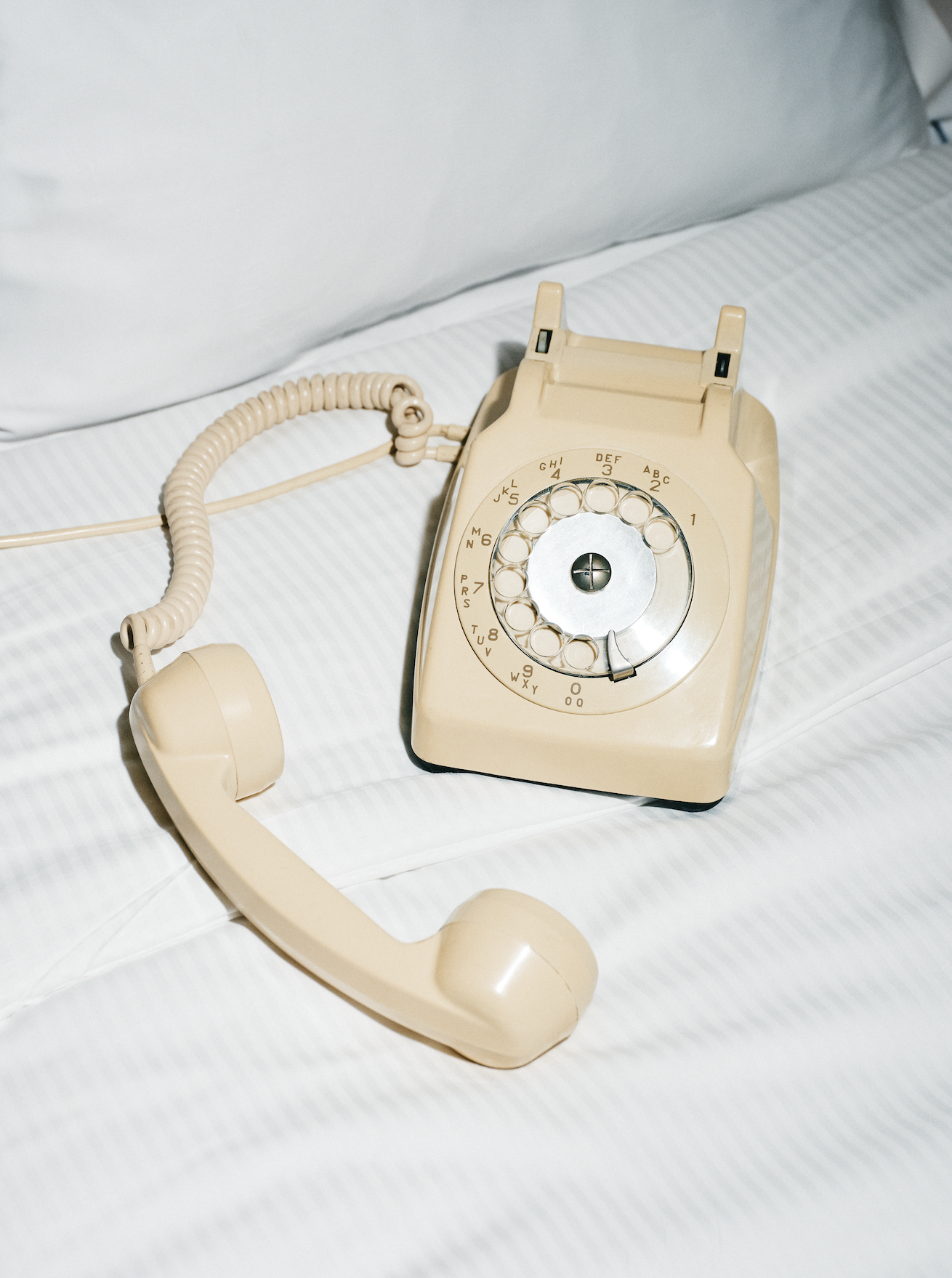 A vintage beige rotary dial telephone resting on a white bed sheet.