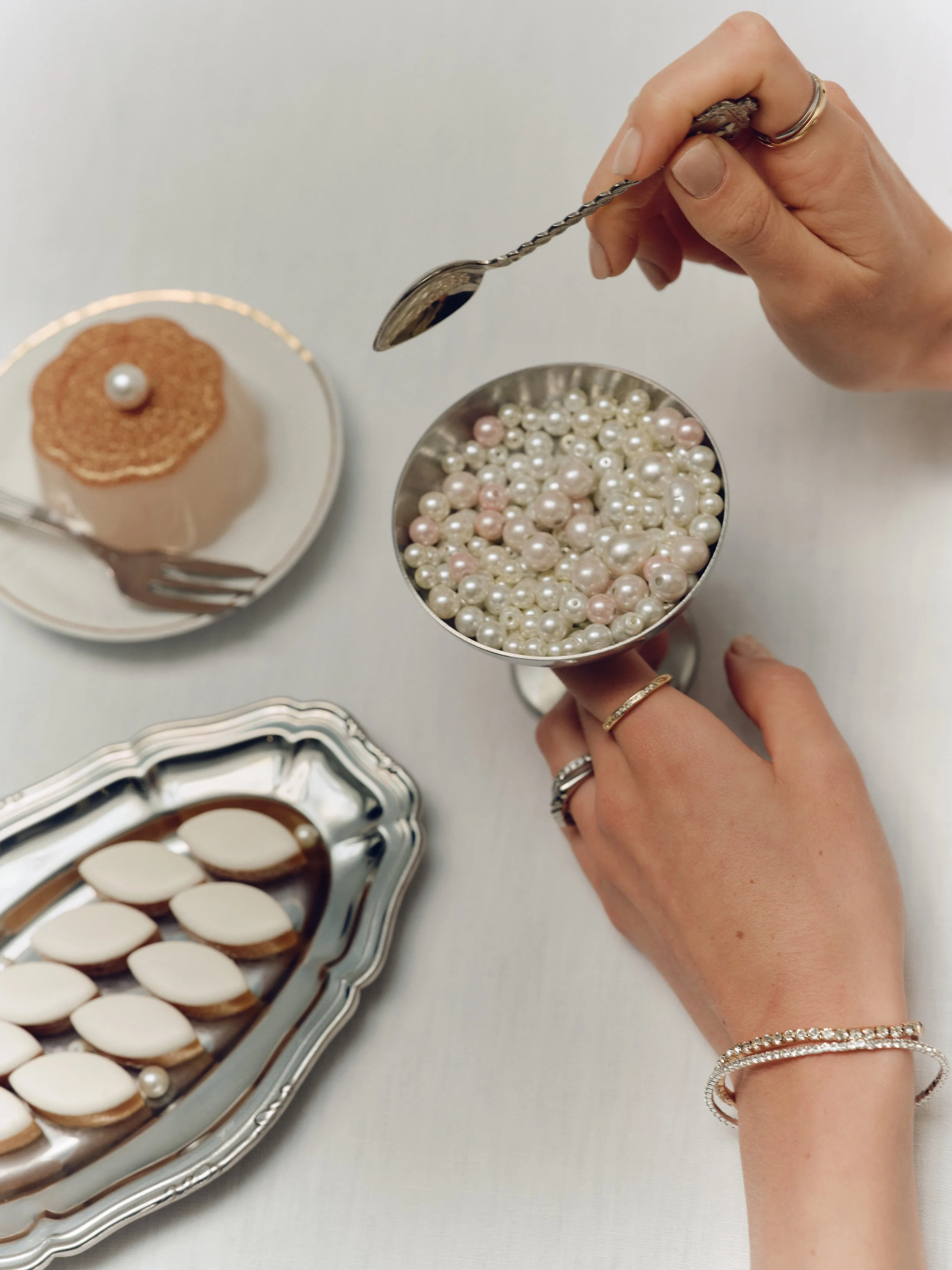 Person holding a silver spoon and wearing rings and bracelets, next to a bowl of pearls and a tray of decorated cookies.
