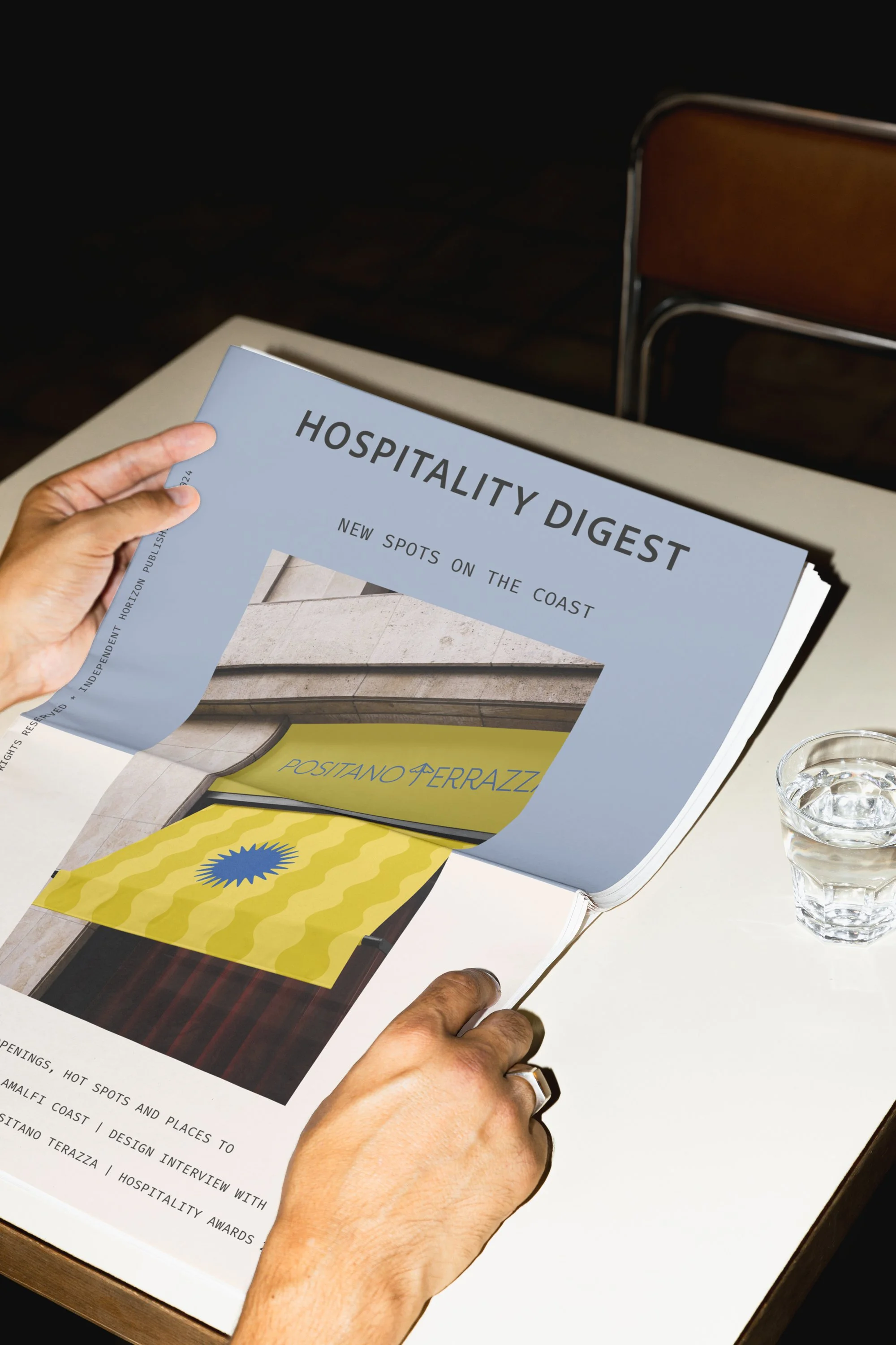 A person holding a magazine titled 'Hospitality Digest' with headlines about new spots on the coast and design interviews, next to a glass of water, with a brown chair in the background.
