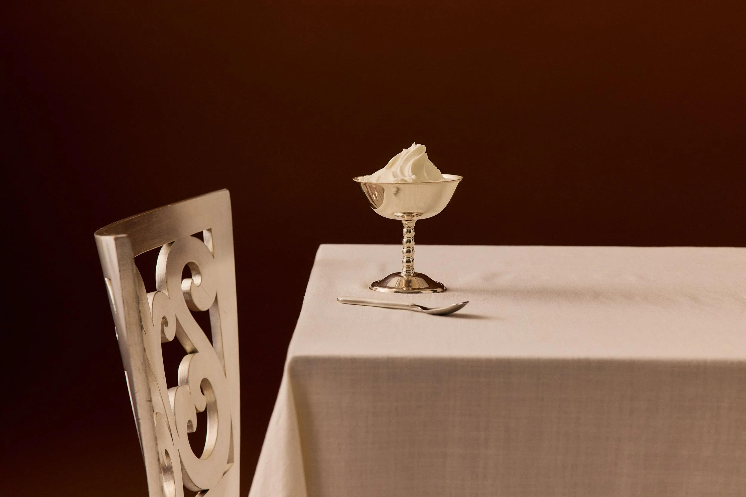 A dining table with a white tablecloth, a silver spoon, a silver dessert dish with whipped cream, and a white chair with a decorative backrest.