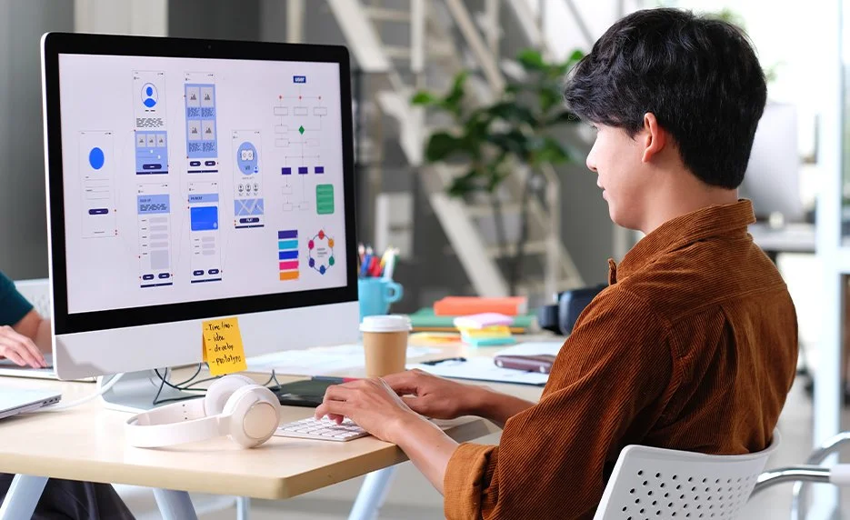 Person working on a desktop computer with wireframe UI designs on the monitor in an office setting.